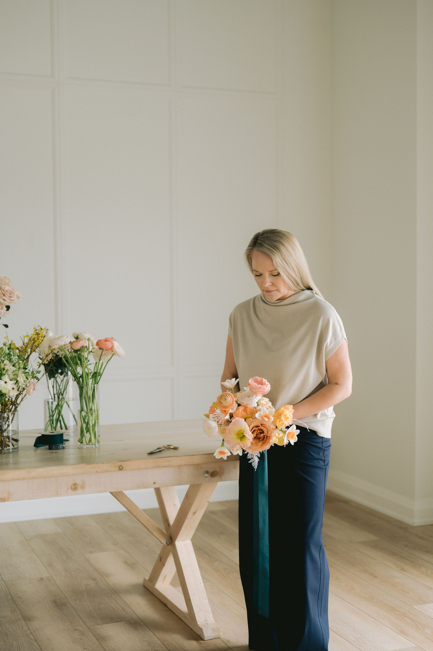 A person arranges peach and coral colored flowers in a bright minimalist interior with wooden floors.