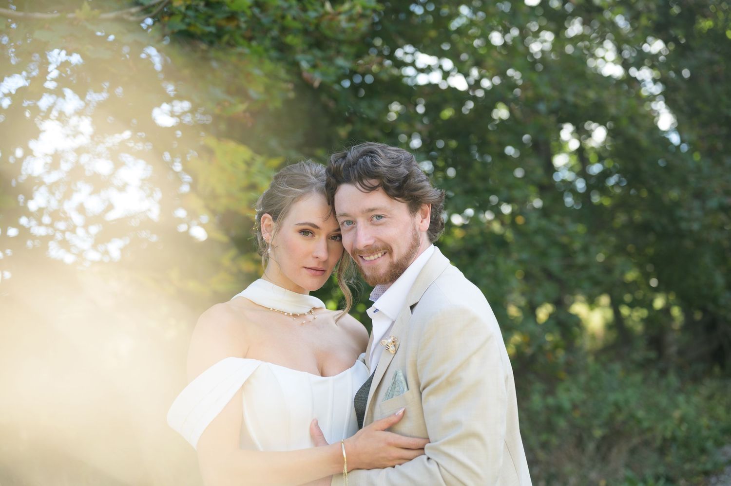 A romantic outdoor wedding portrait of a couple embracing in soft sunlight with trees in the background.