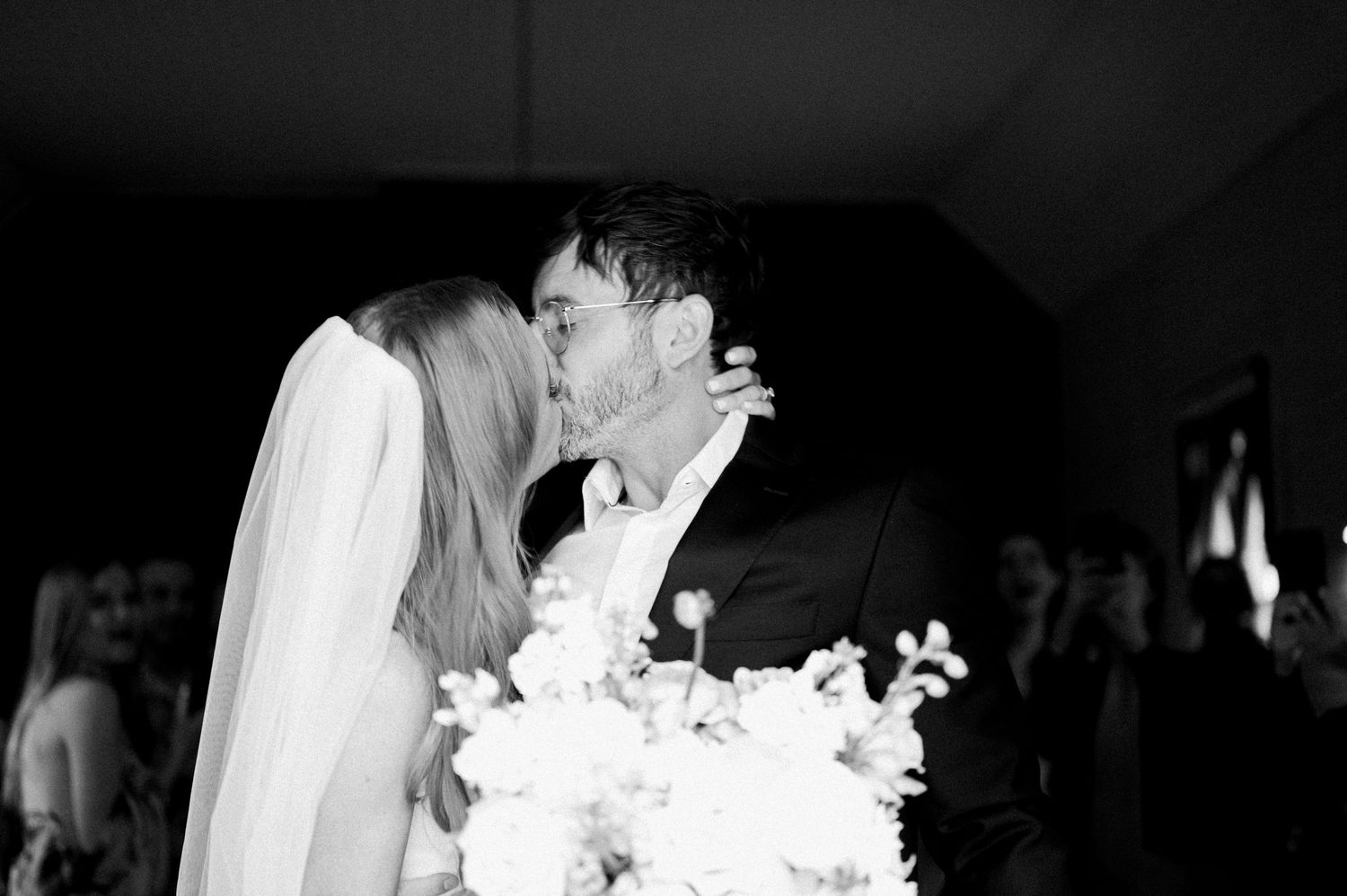 A black and white wedding photograph showing a romantic kiss between newlyweds holding a white floral bouquet.