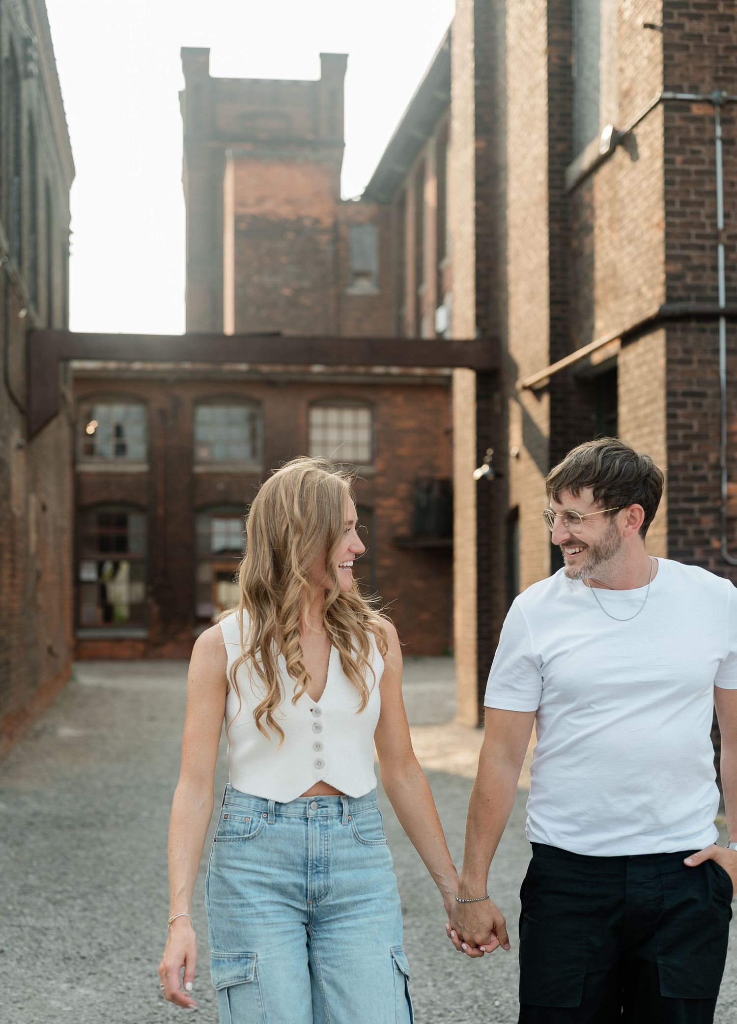 Couple in casual white tops and jeans holding hands while walking through an industrial brick building setting.