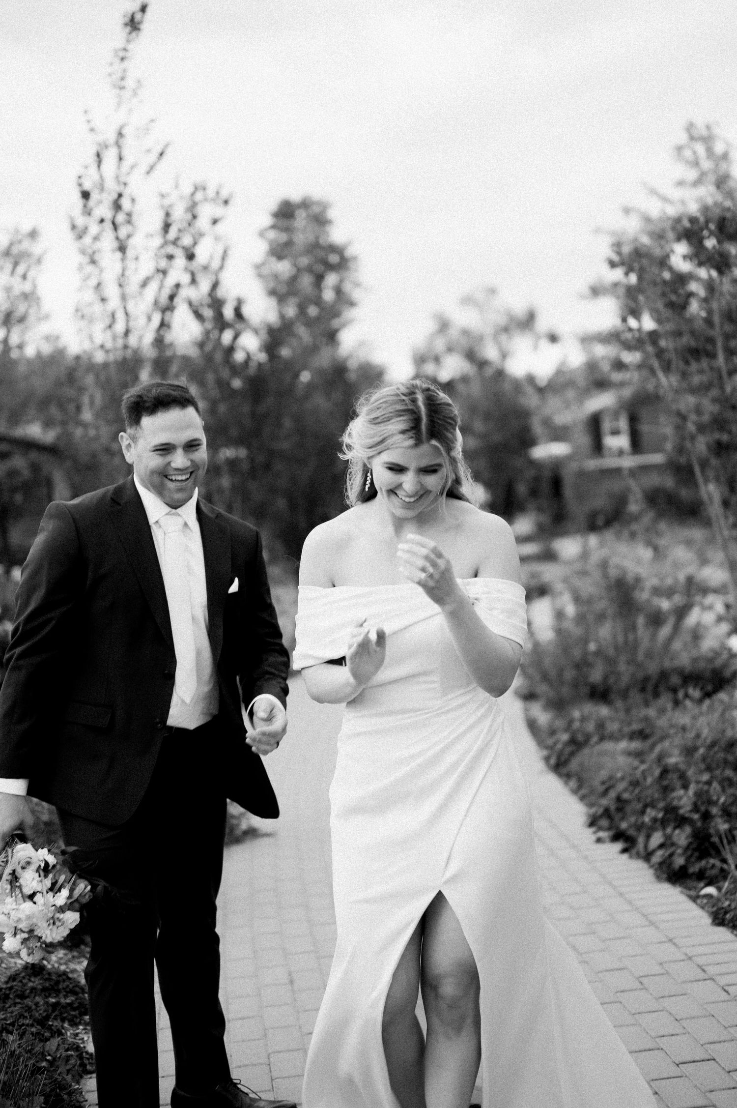 A happy couple shares joyful moments on their wedding day walking along a stone path in black and white photography.