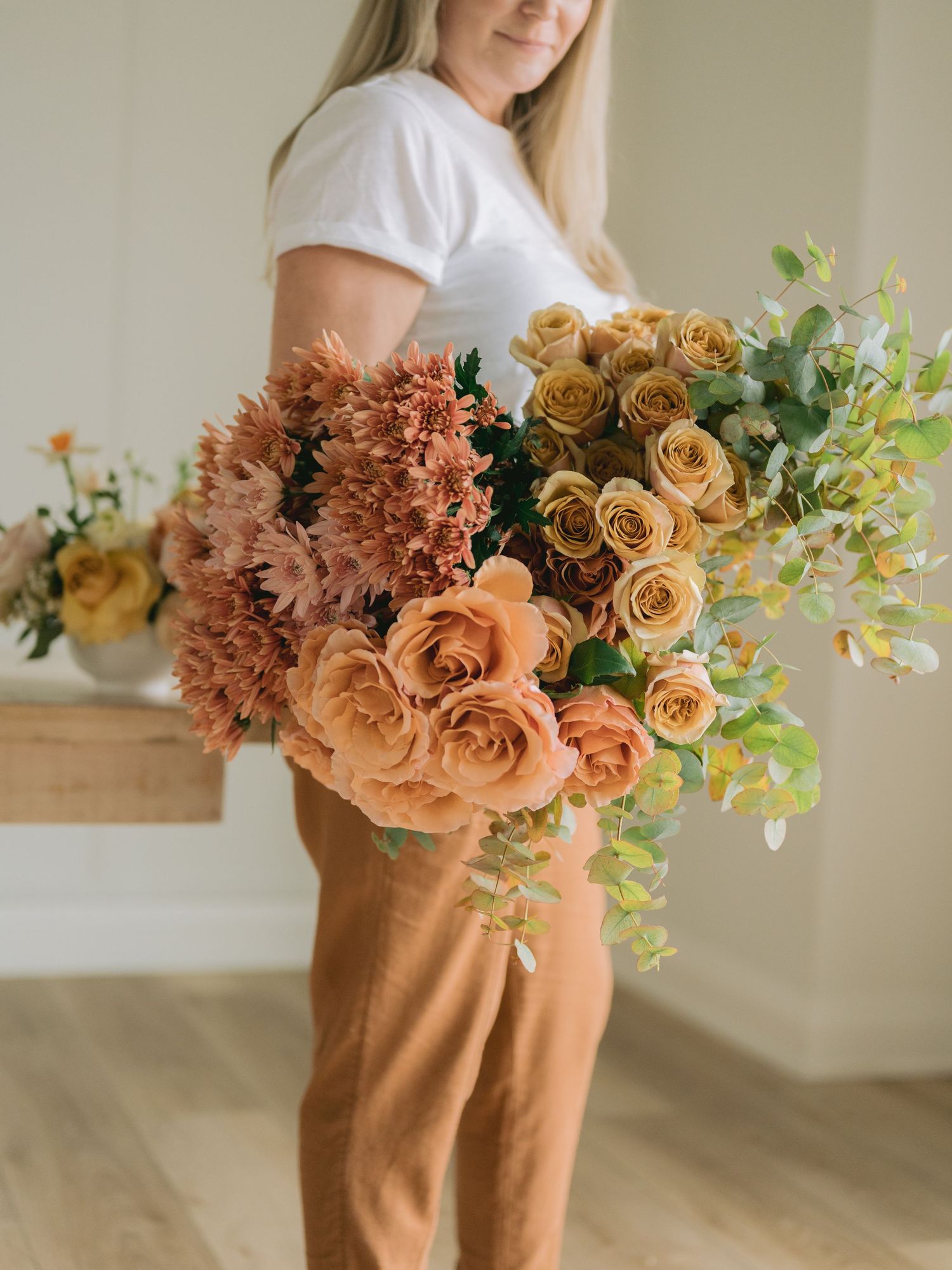 Someone in white shirt and tan pants holding a lush bouquet of peach roses, chrysanthemums and greenery.
