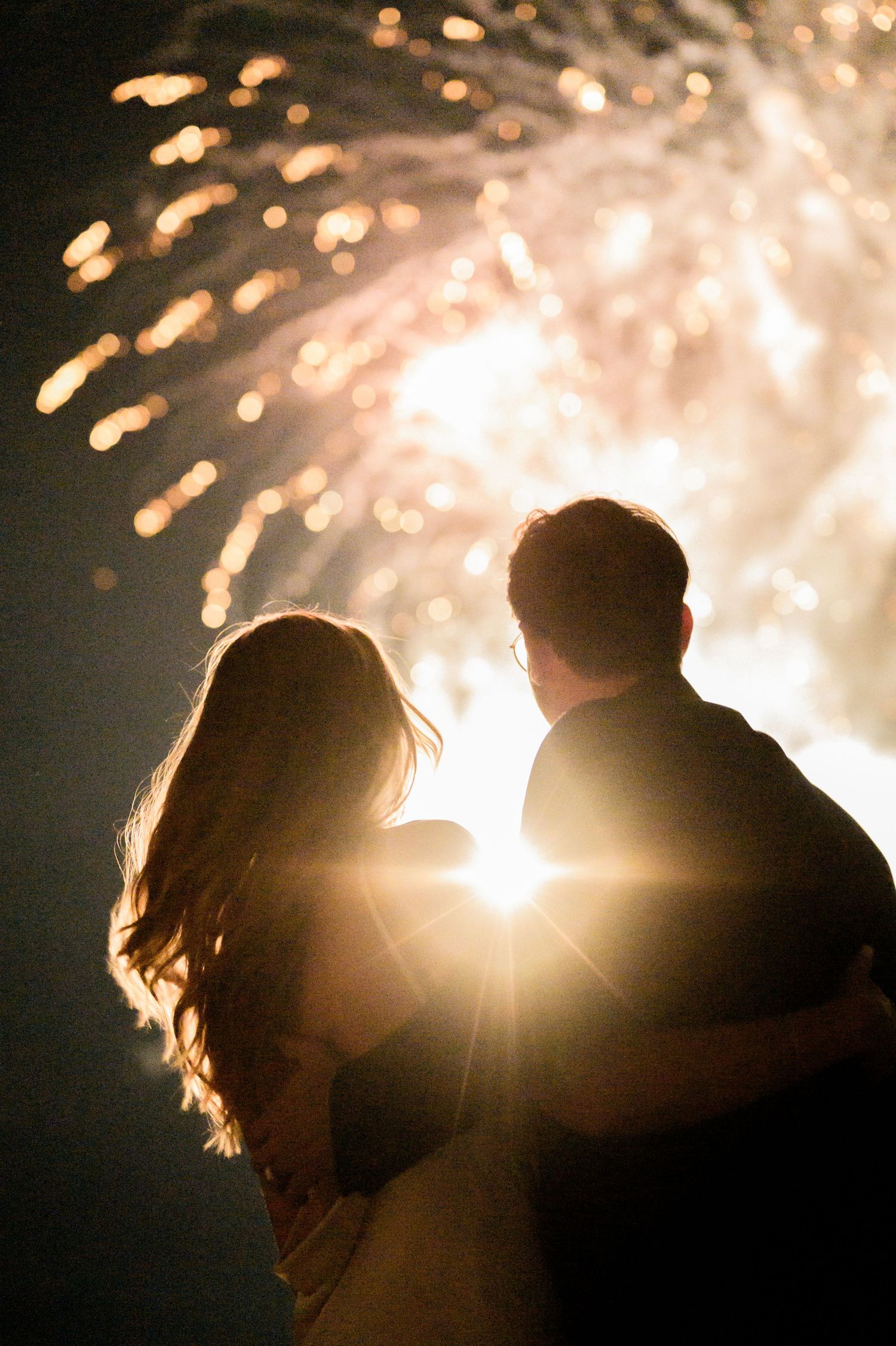 A silhouetted couple watches fireworks together against a dark sky with glowing sparks and bokeh lights.
