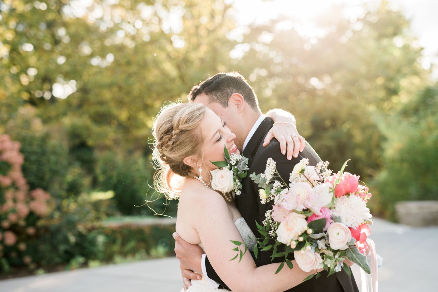 Romantic couple shares tender kiss in golden sunlit garden setting with pink and white flowers in the background.
