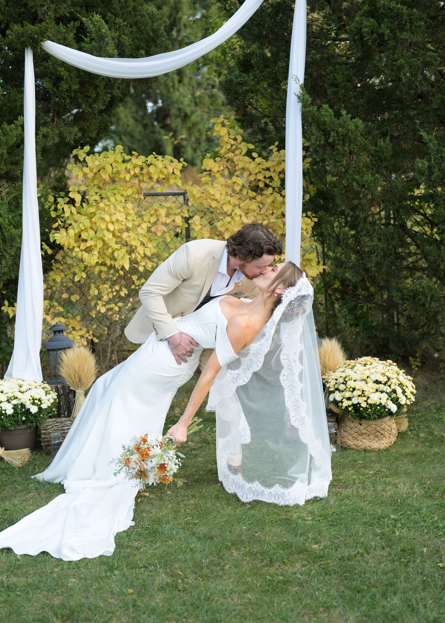 A playful wedding couple poses behind flowing white fabric drapes in an outdoor garden setting.
