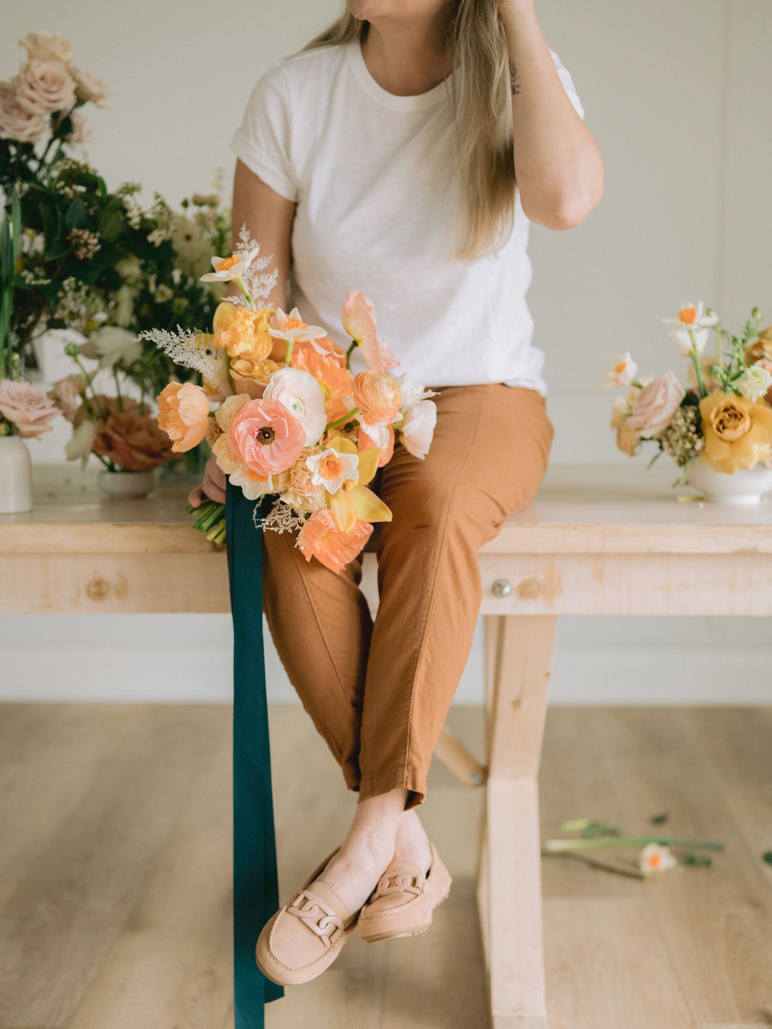 A person in a white t-shirt and tan pants sits casually on a wooden bench with peach-colored flowers nearby.
