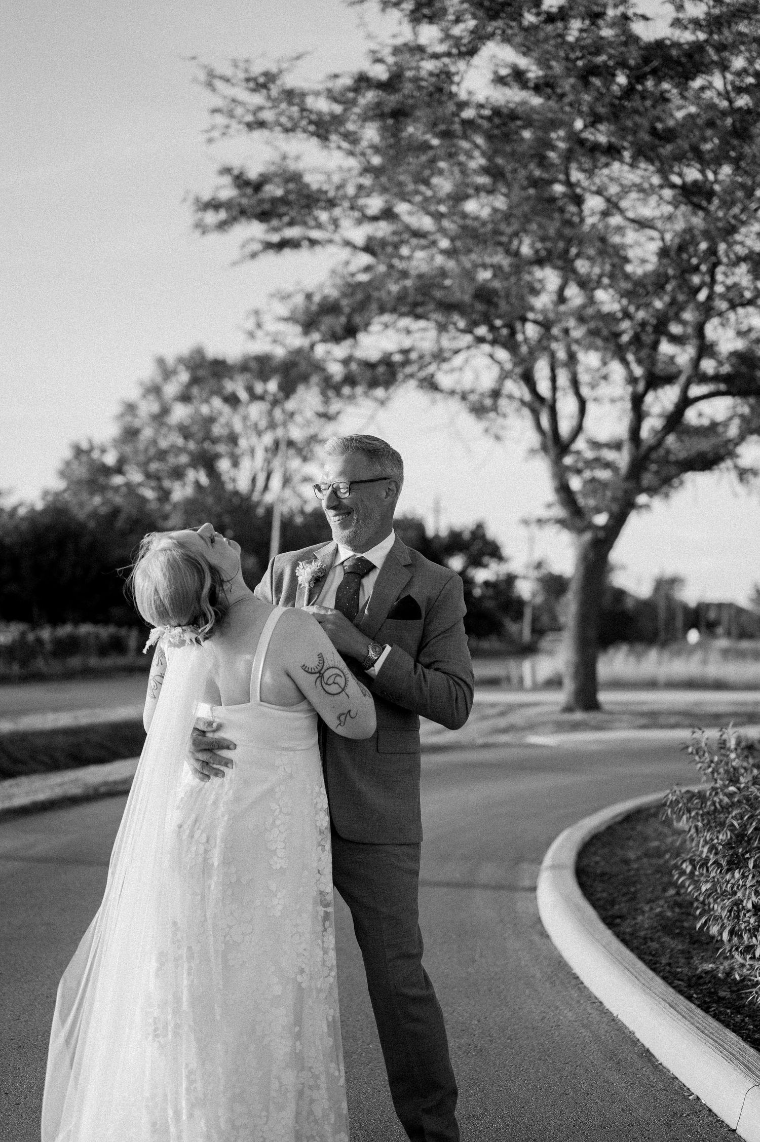 A couple in wedding attire dances together on a tree-lined path in a romantic black and white photo series.
