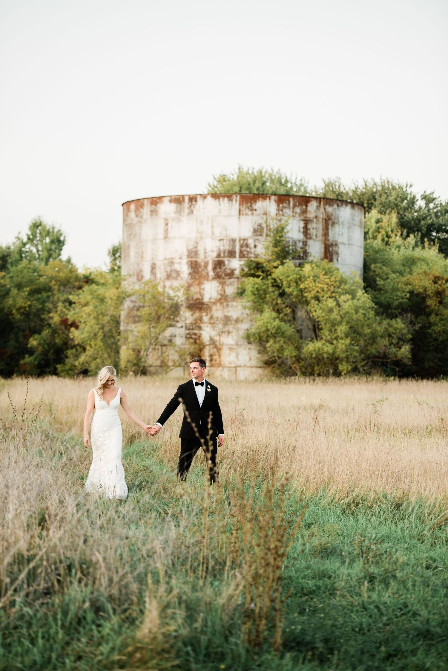 A couple in wedding attire walks through a rustic field with tall grass and industrial buildings in the background.