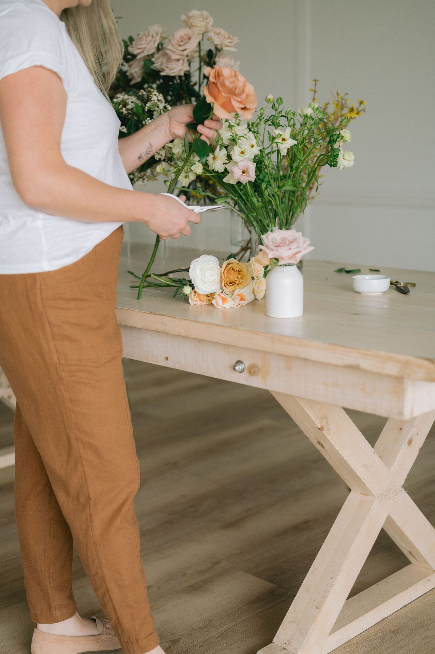 Person arranging fresh flowers in white vases on a rustic wooden table wearing brown pants and white top.