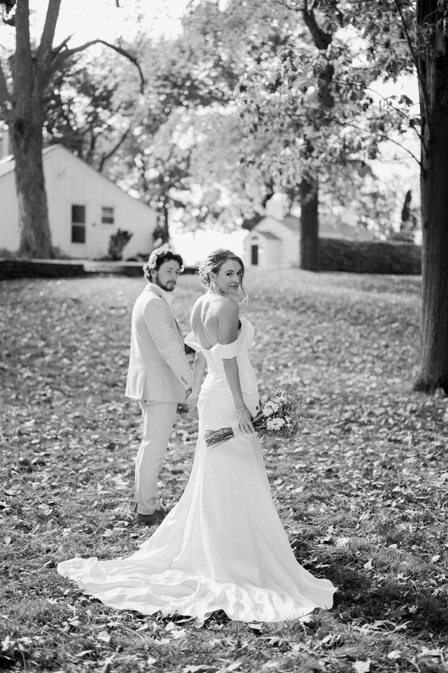 A black and white wedding photo showing a couple in formal attire standing among autumn leaves on a lawn.