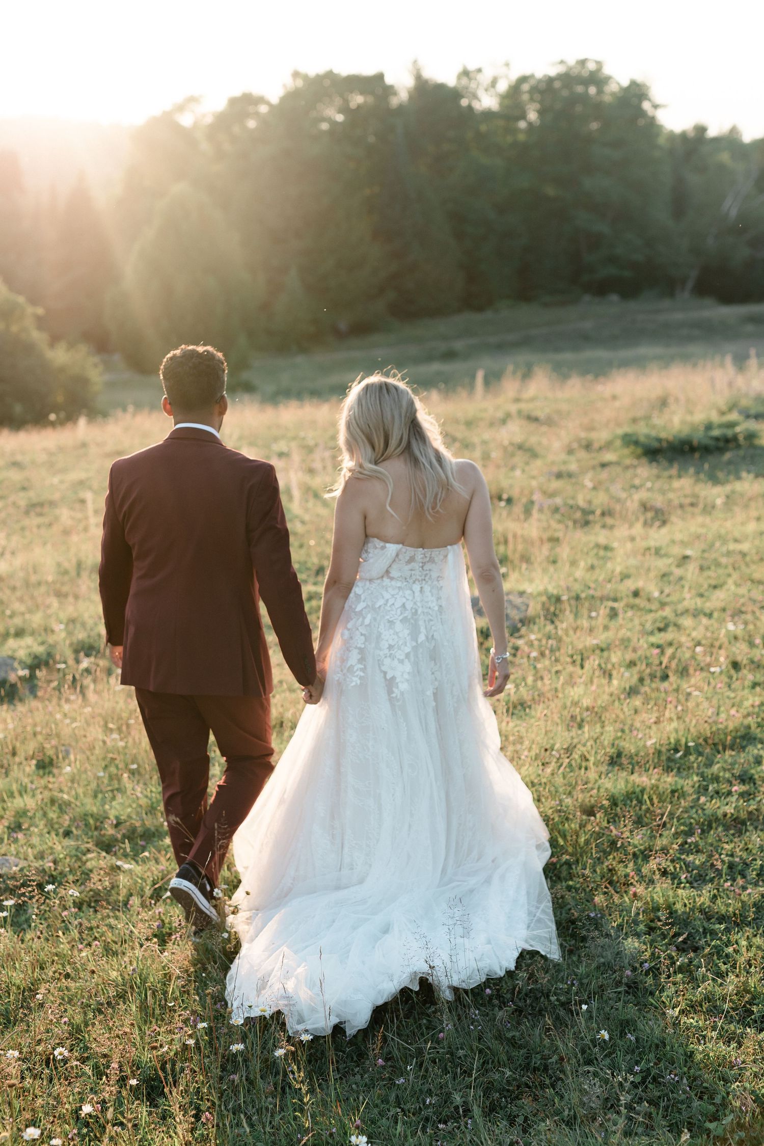 Newlywed couple walking hand in hand through a sunlit meadow at golden hour, bride wearing flowing lace gown.