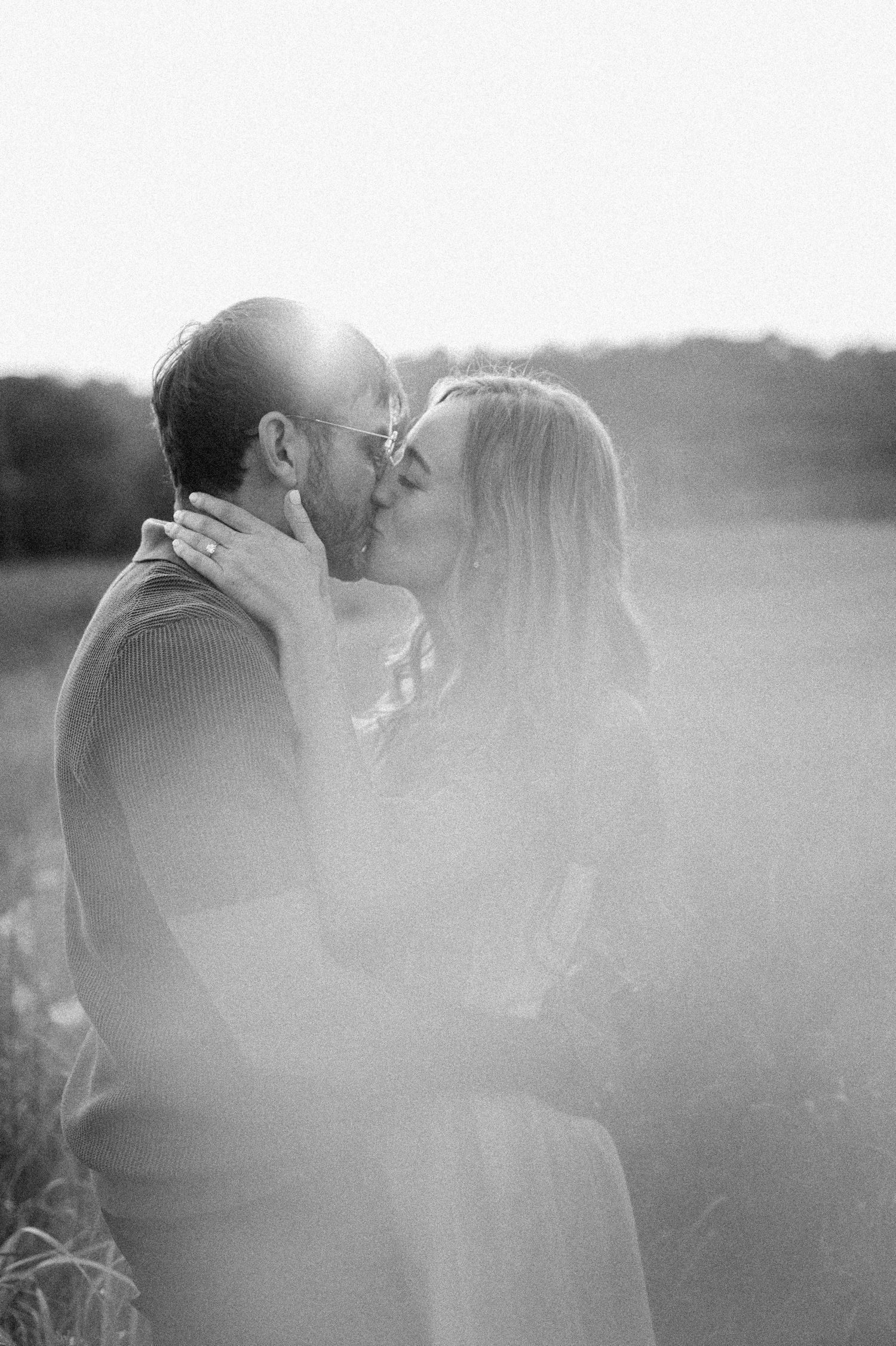 A romantic black and white photo series showing a couple sharing tender kisses in a misty outdoor setting at sunset.
