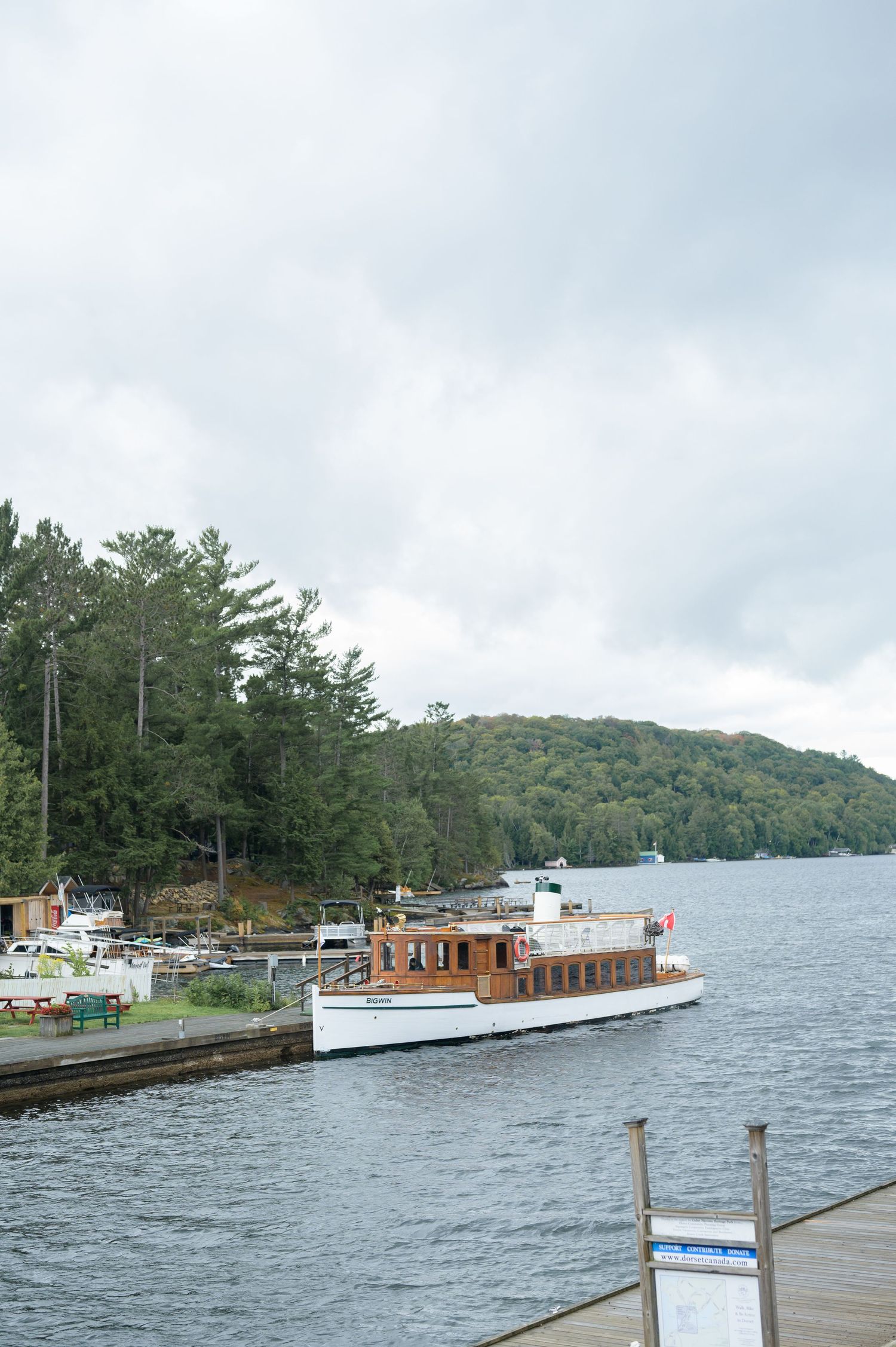 A classic wooden tour boat docked at a lakeside pier surrounded by pine trees and rolling hills under cloudy skies.