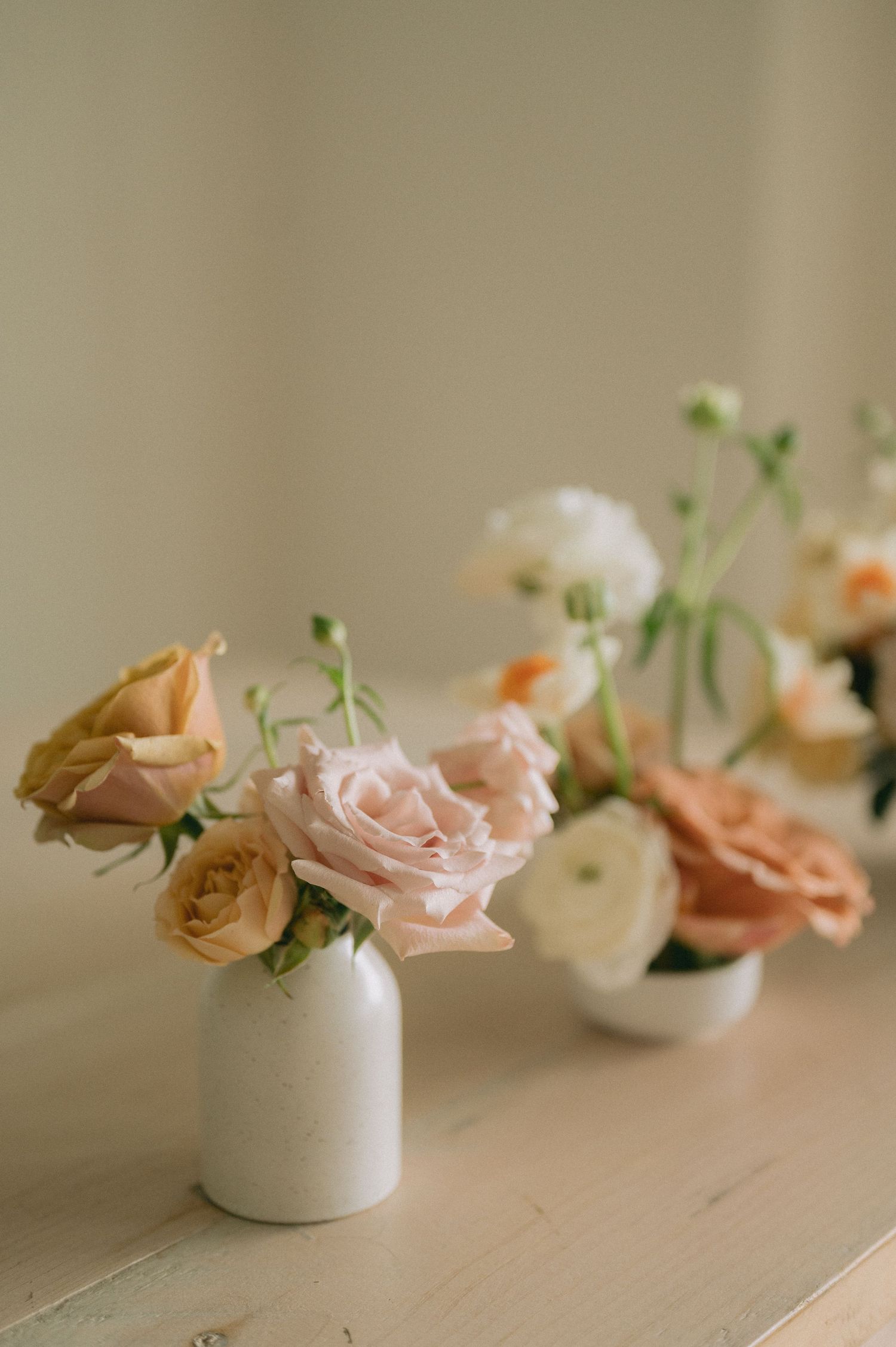 Delicate pink roses and small white flowers arranged in petite white ceramic vases on a light wooden surface.
