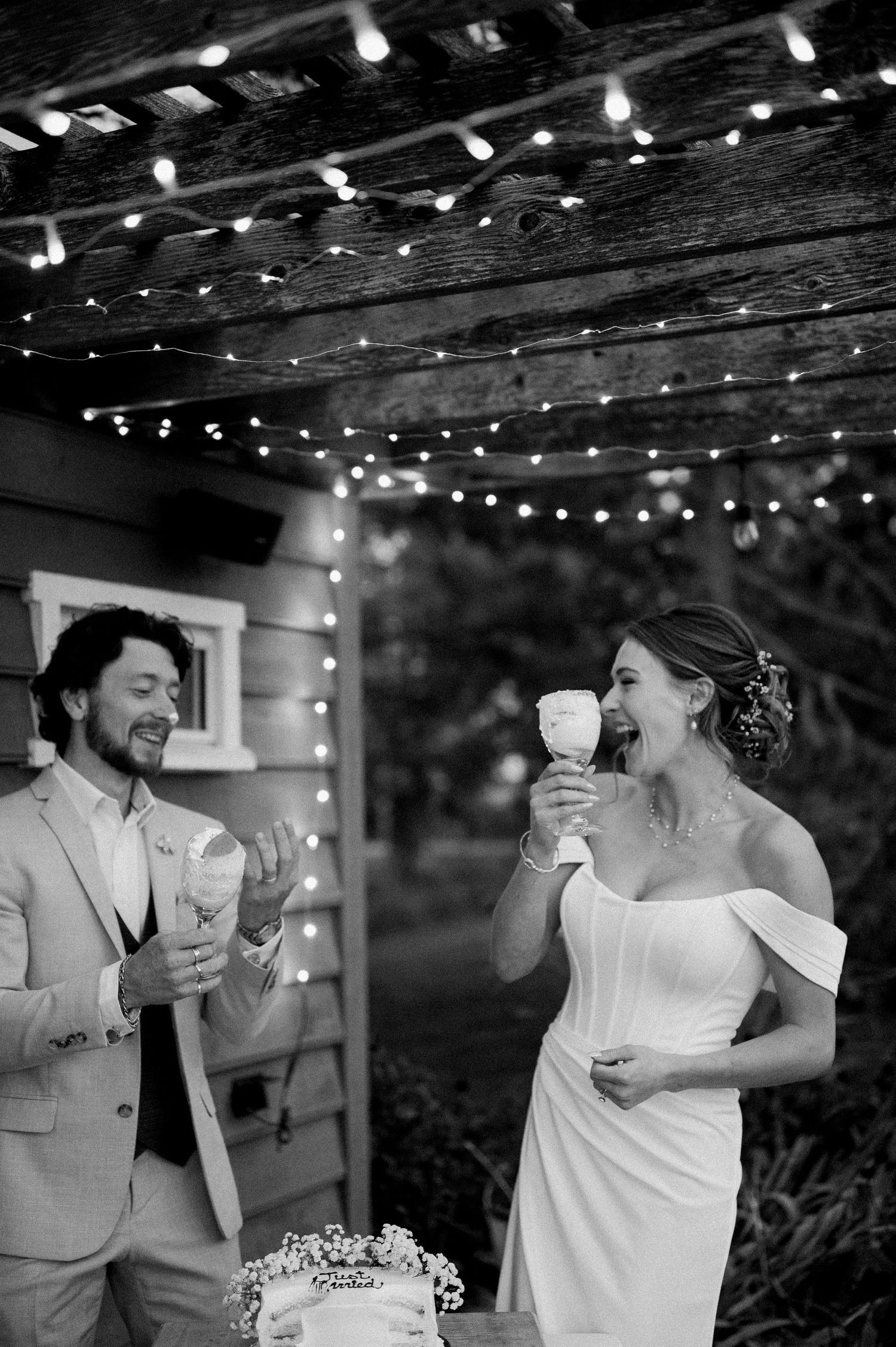 Black and white photo of a bride and groom sharing cake cutting wine glasses under string lights at an outdoor wedding reception in Milton.