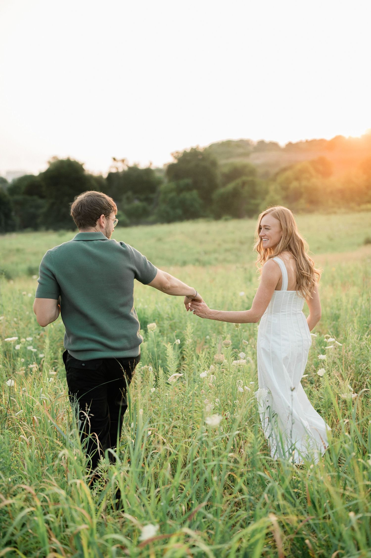 A couple holds hands while running through a sunlit meadow at golden hour, their figures silhouetted against tall grass.