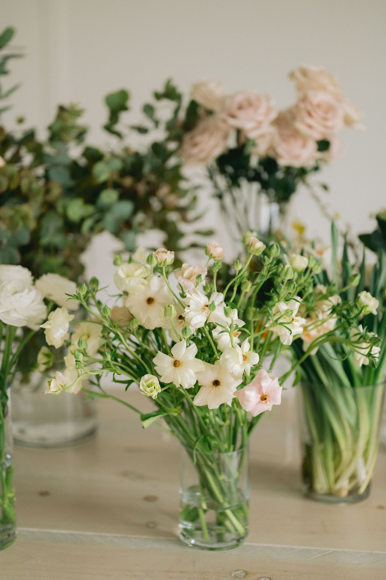 A row of glass vases filled with white and pink flowers and greenery arranged on a light surface.