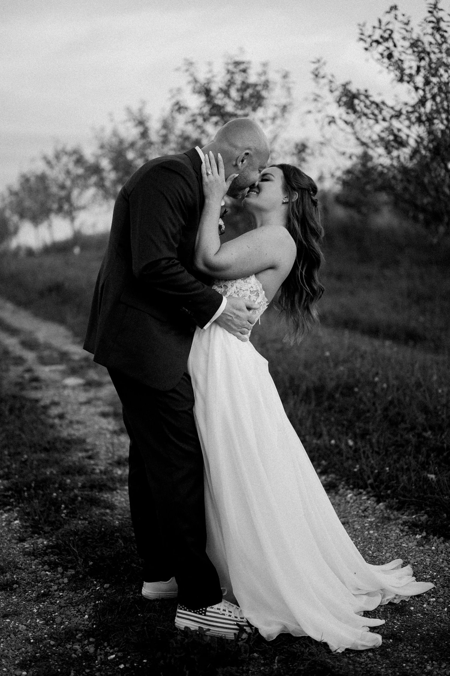 A romantic black and white wedding photo shows a couple kissing on a country path with trees in the background.