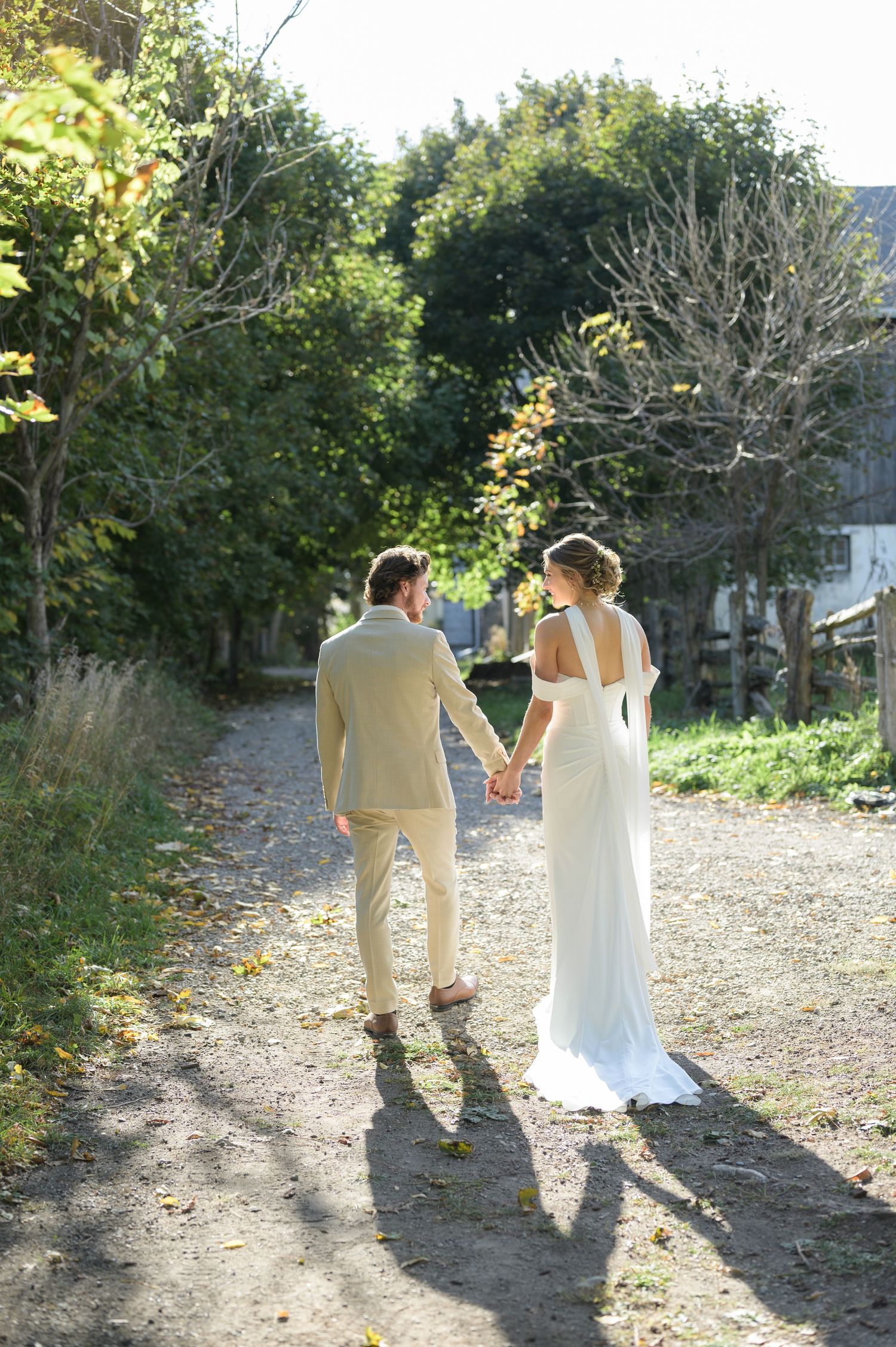 A couple walking away together down a sunlit garden path in wedding attire.