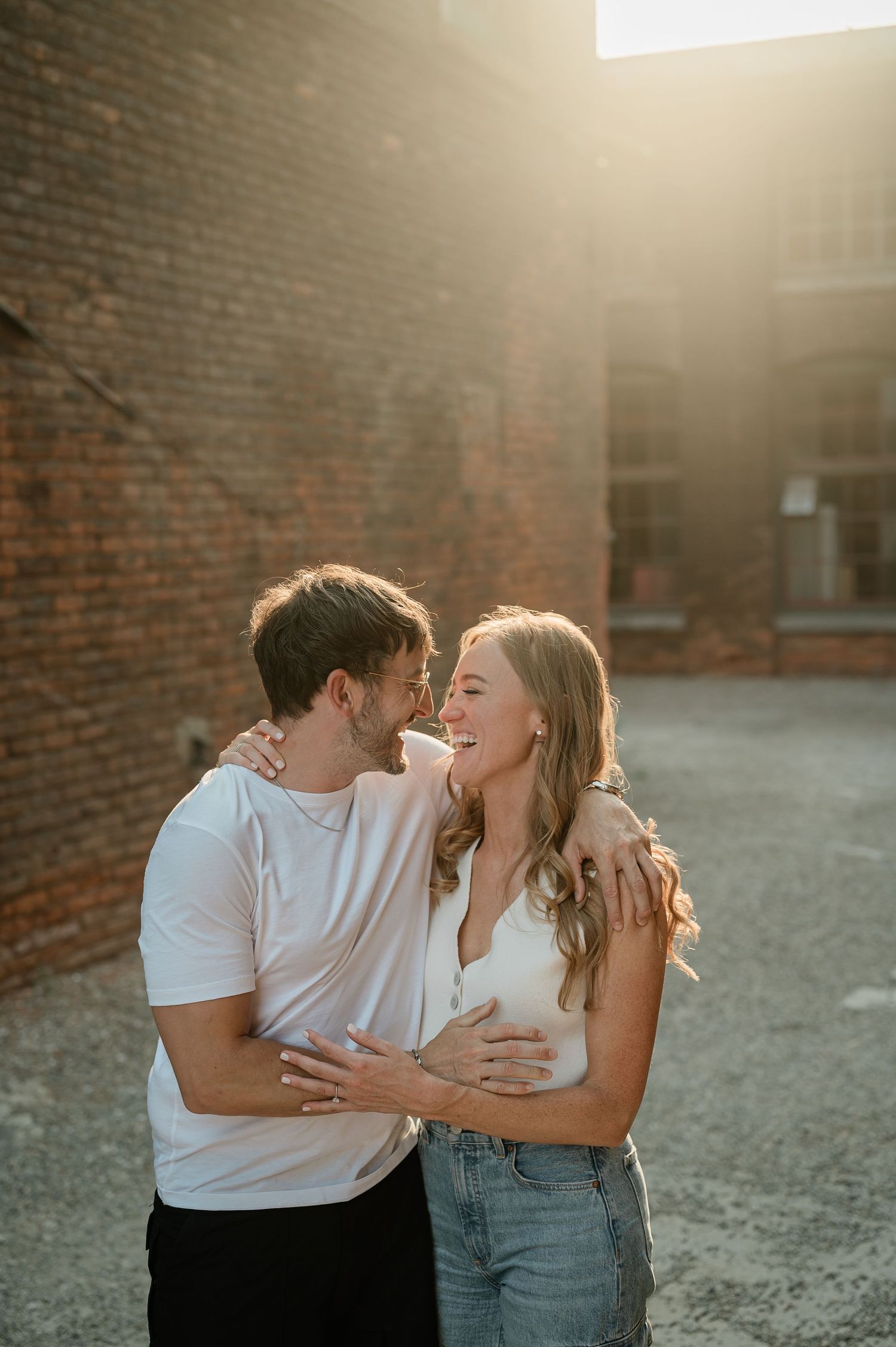 A couple shares an intimate moment together while embracing against a brick wall in warm sunset lighting.