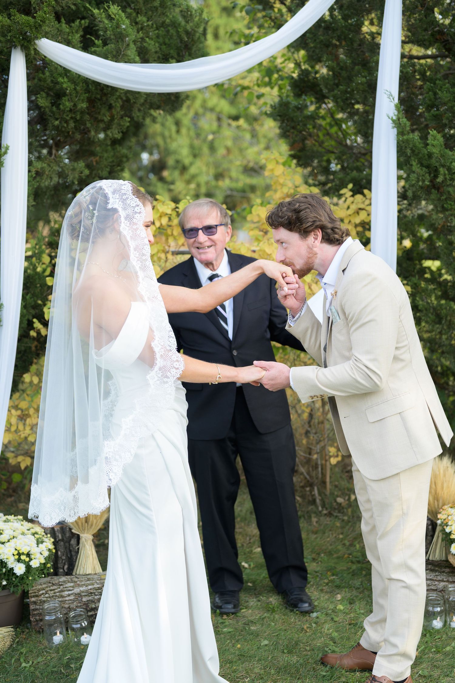 An outdoor wedding ceremony under flowing white fabric draped between trees in a garden setting.