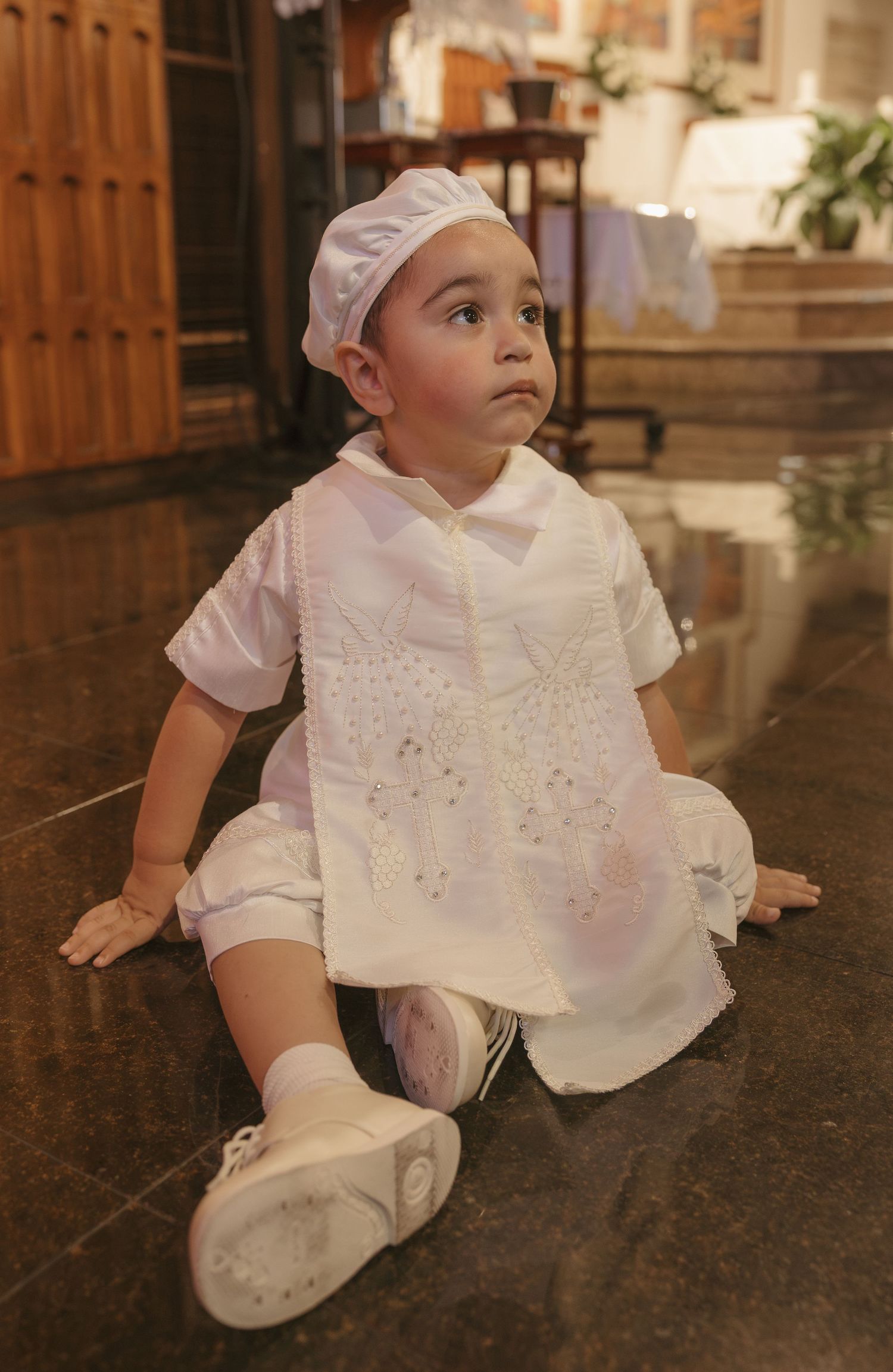 Baby in white christening outfit with embroidered bib and matching hat sits on marble floor.