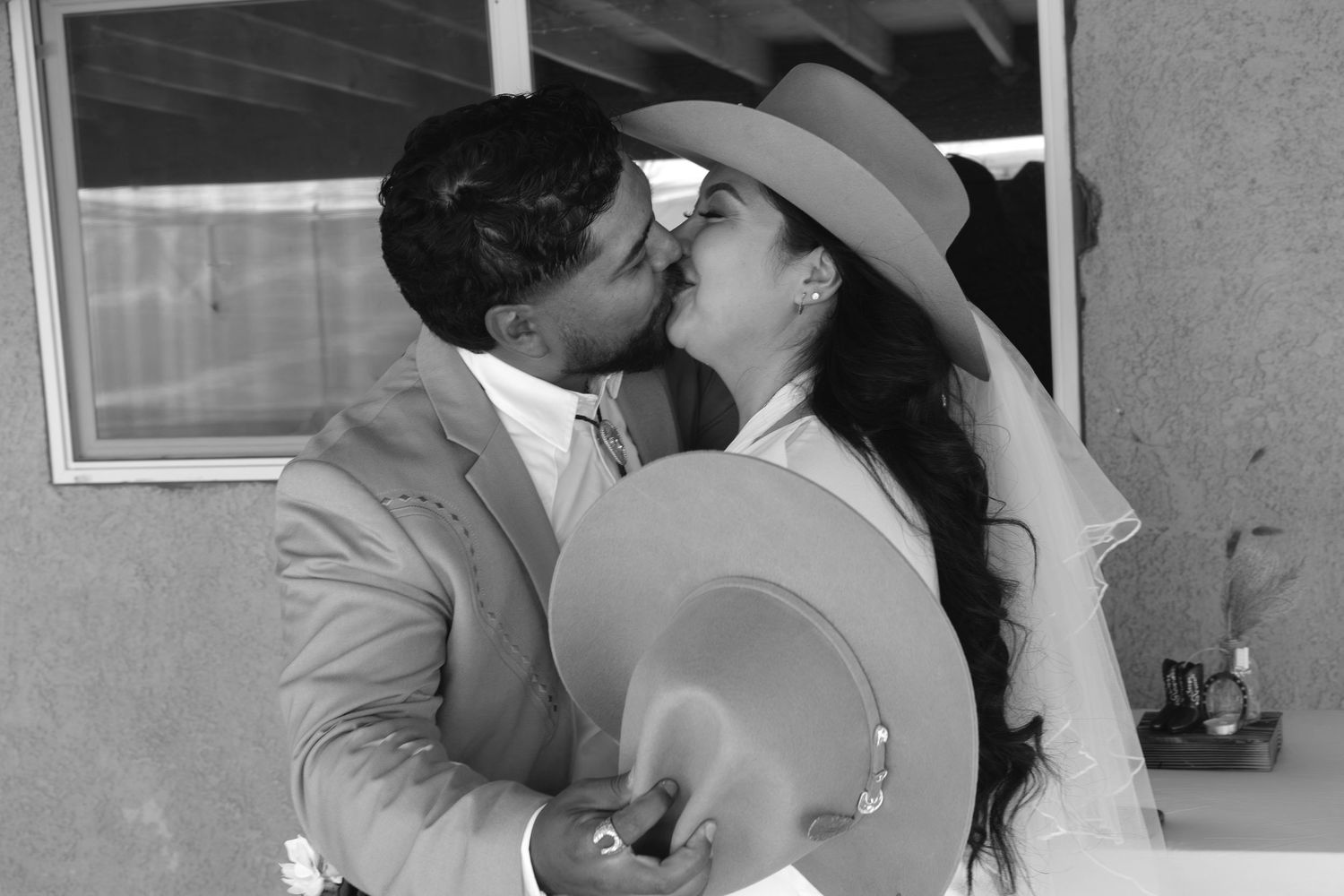 Black and white photo of a couple sharing a romantic kiss, bride wearing a wide-brim hat at their wedding.