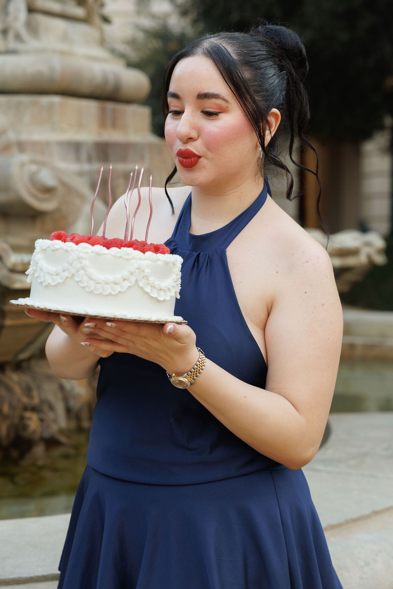 Young woman in navy dress blowing out candles on white birthday cake decorated with raspberries outdoors.