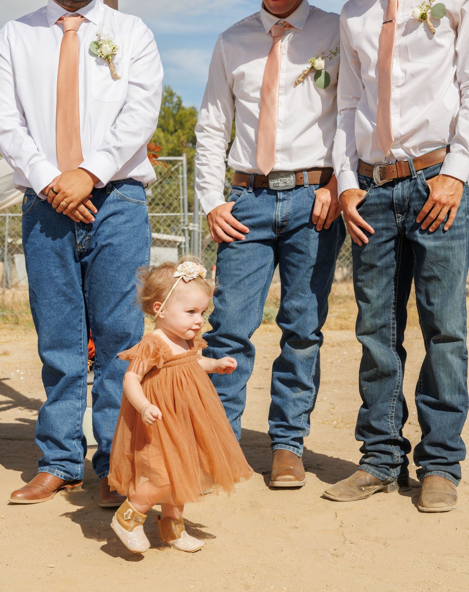Toddler flower girl in rust tulle dress walking in front of groomsmen in jeans and pink ties.