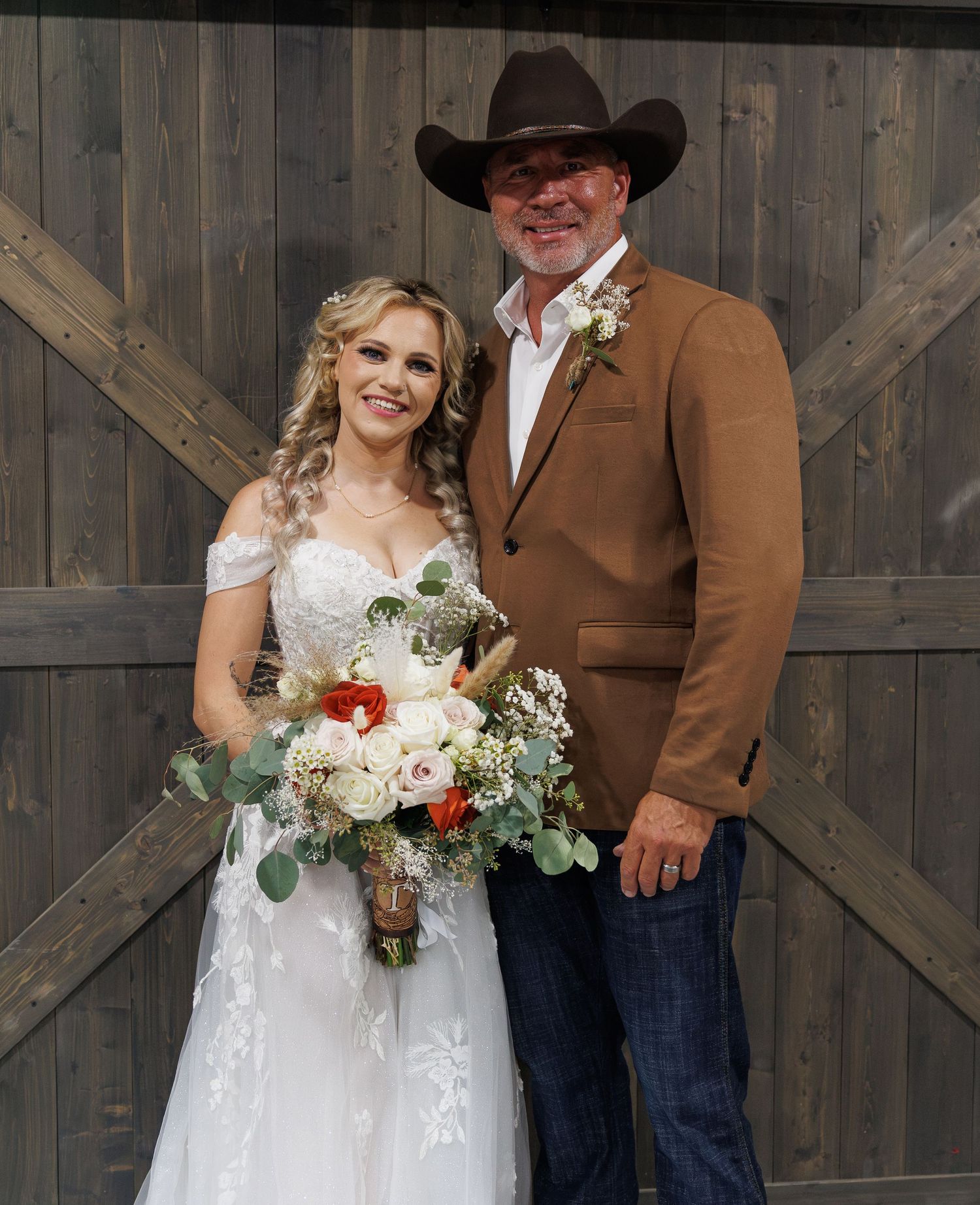 Bride in white lace gown holding floral bouquet stands with groom in brown blazer and cowboy hat before rustic barn doors.