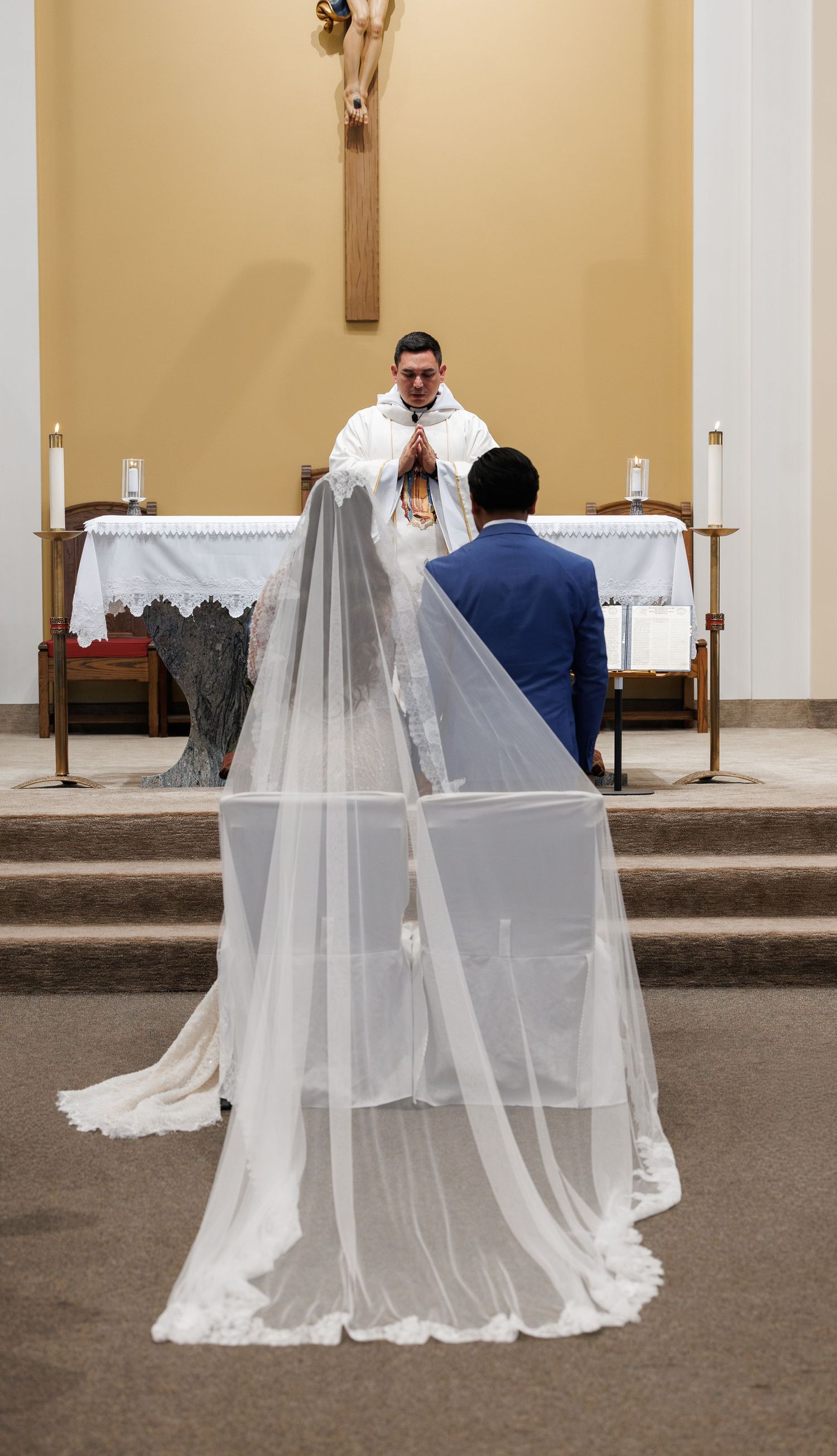 Bride in a long lace-trimmed veil and groom in a blue suit kneel before a priest at a church altar during a wedding ceremony.