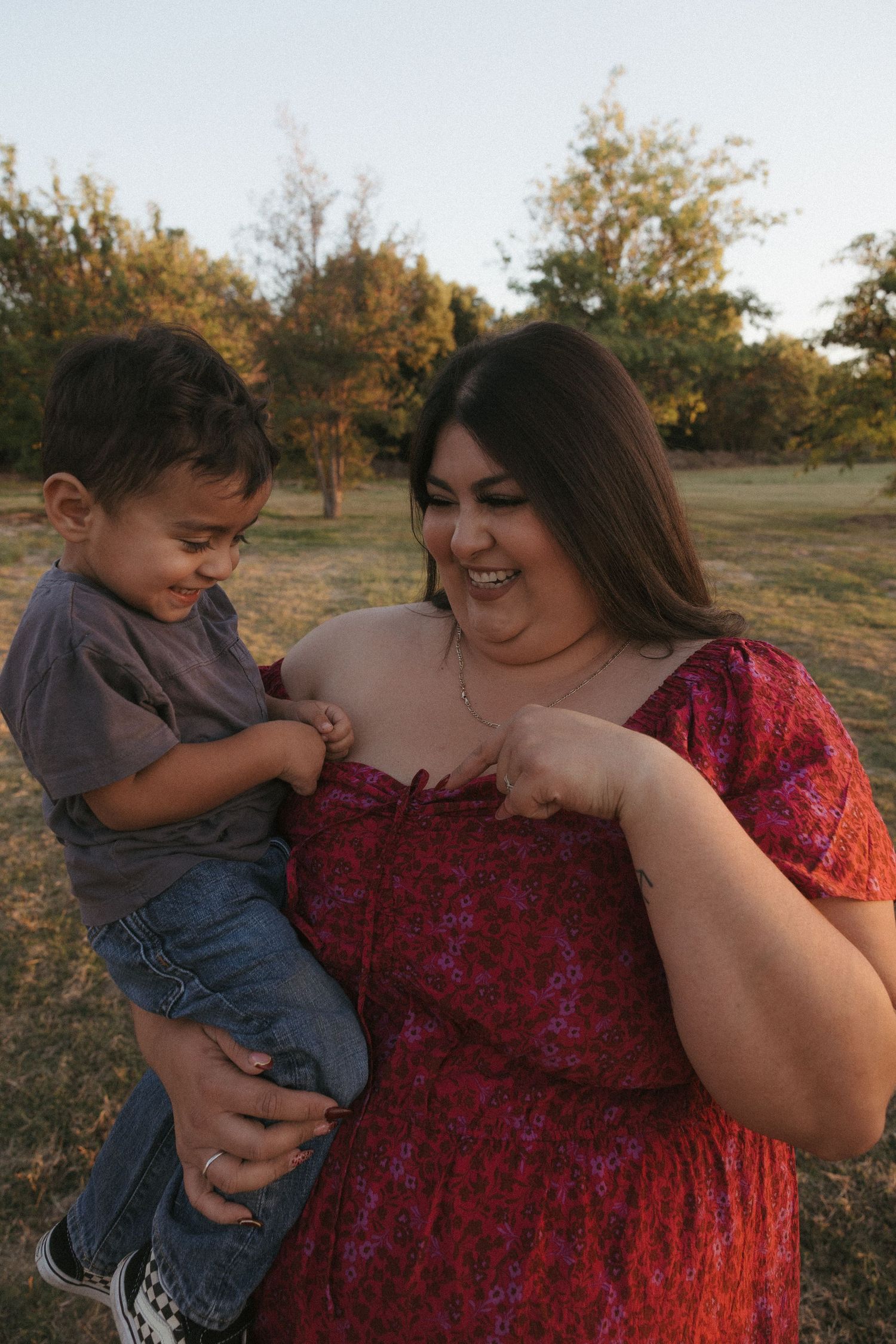 A smiling woman in a red floral dress holds a toddler boy outdoors in a sunny park setting.