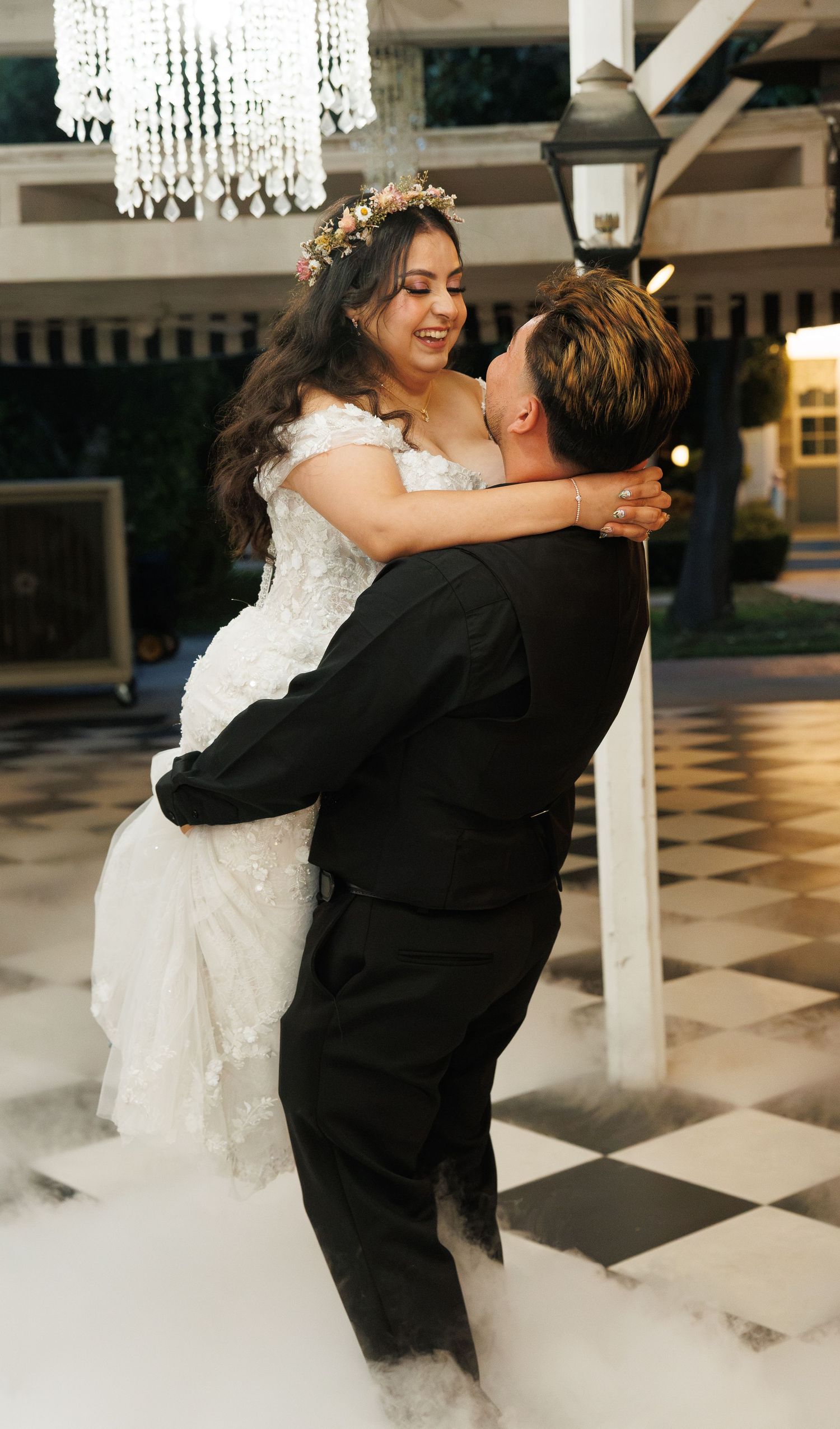 Groom lifting bride in wedding dress on black and white dance floor with fog effects and chandelier.