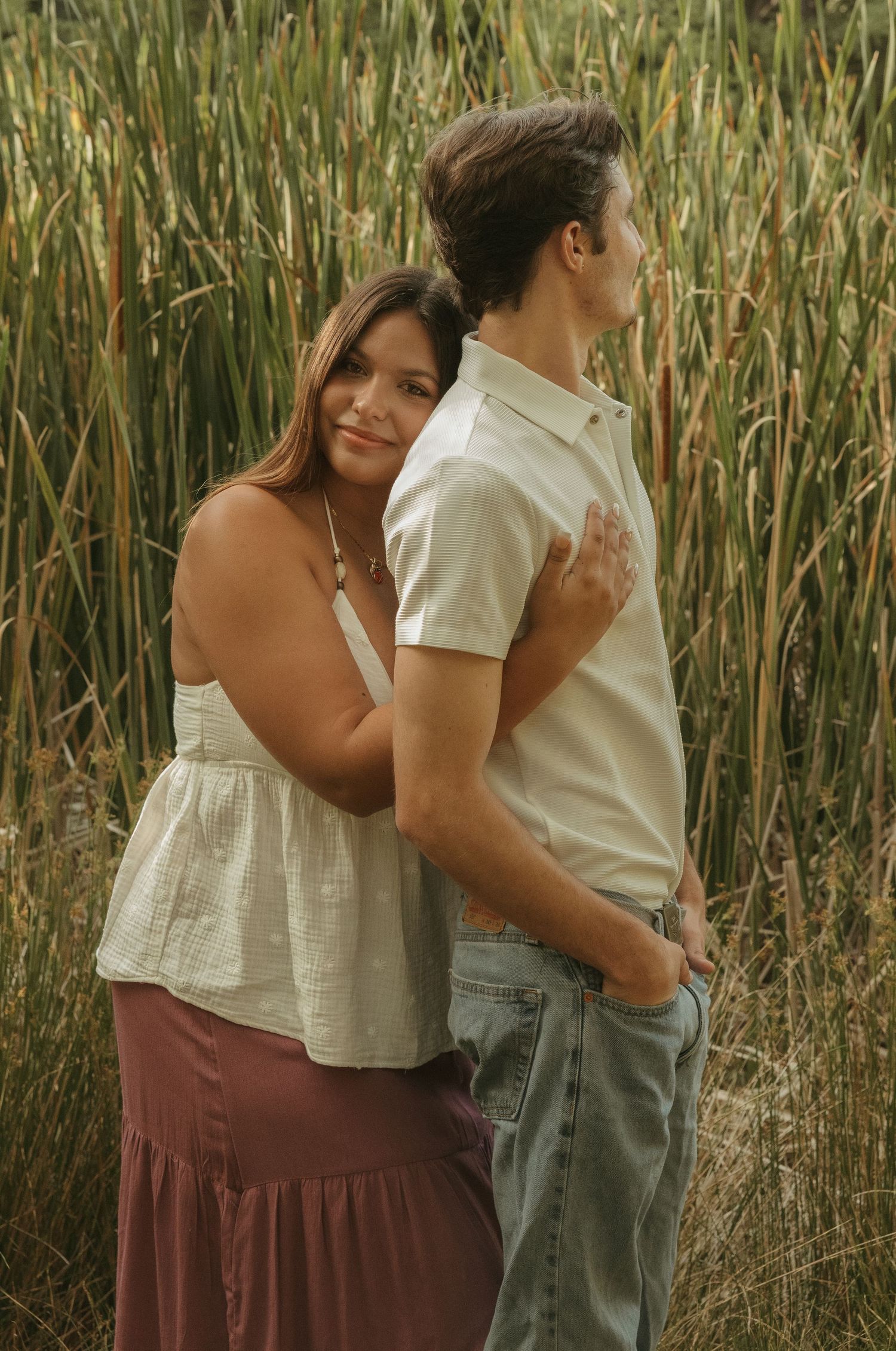 A couple embraces in a field of tall grass, woman in white top and mauve skirt rests her hand on mans back.