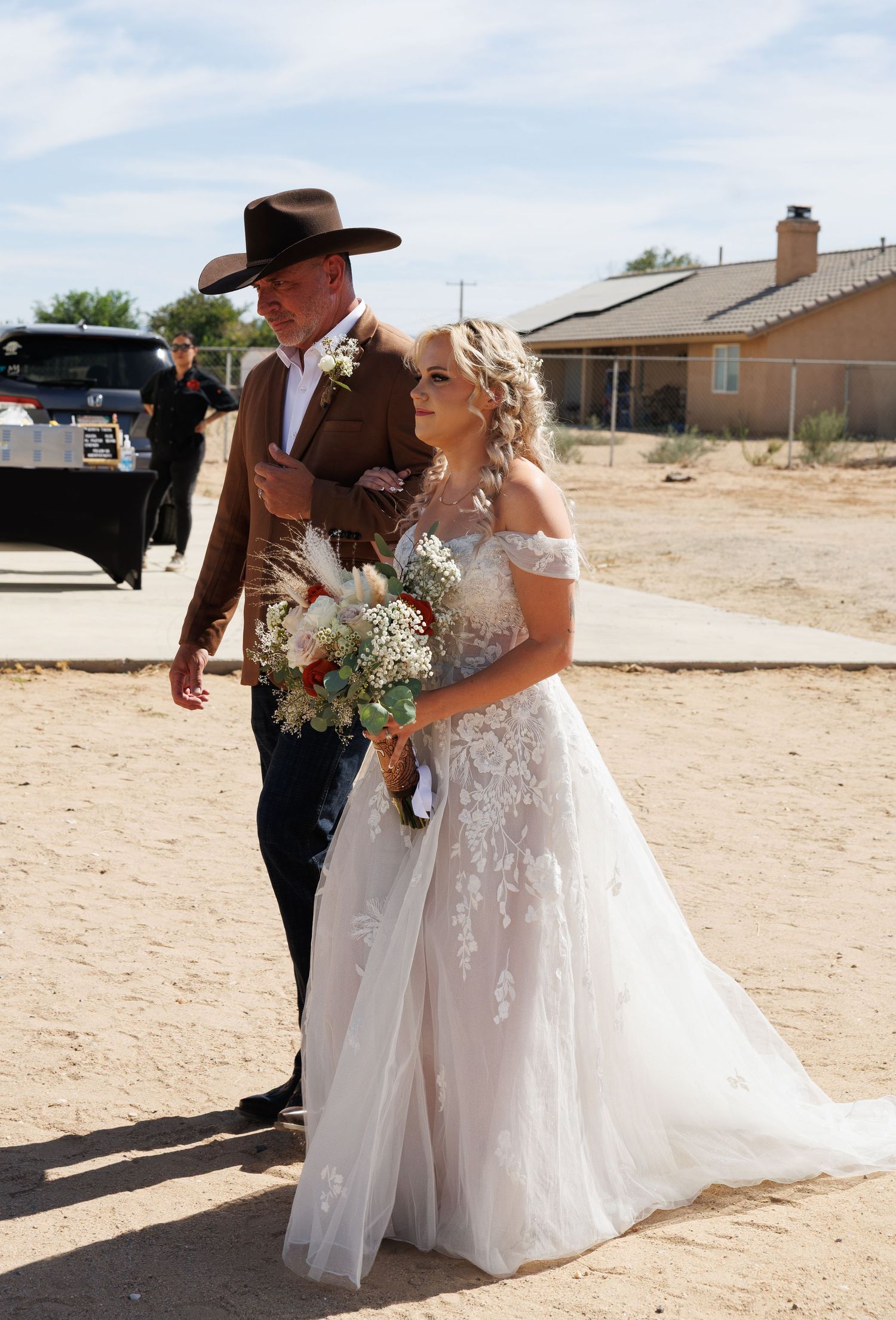Bride in lace off-shoulder gown walks with groom in brown suit and cowboy hat at outdoor western wedding.