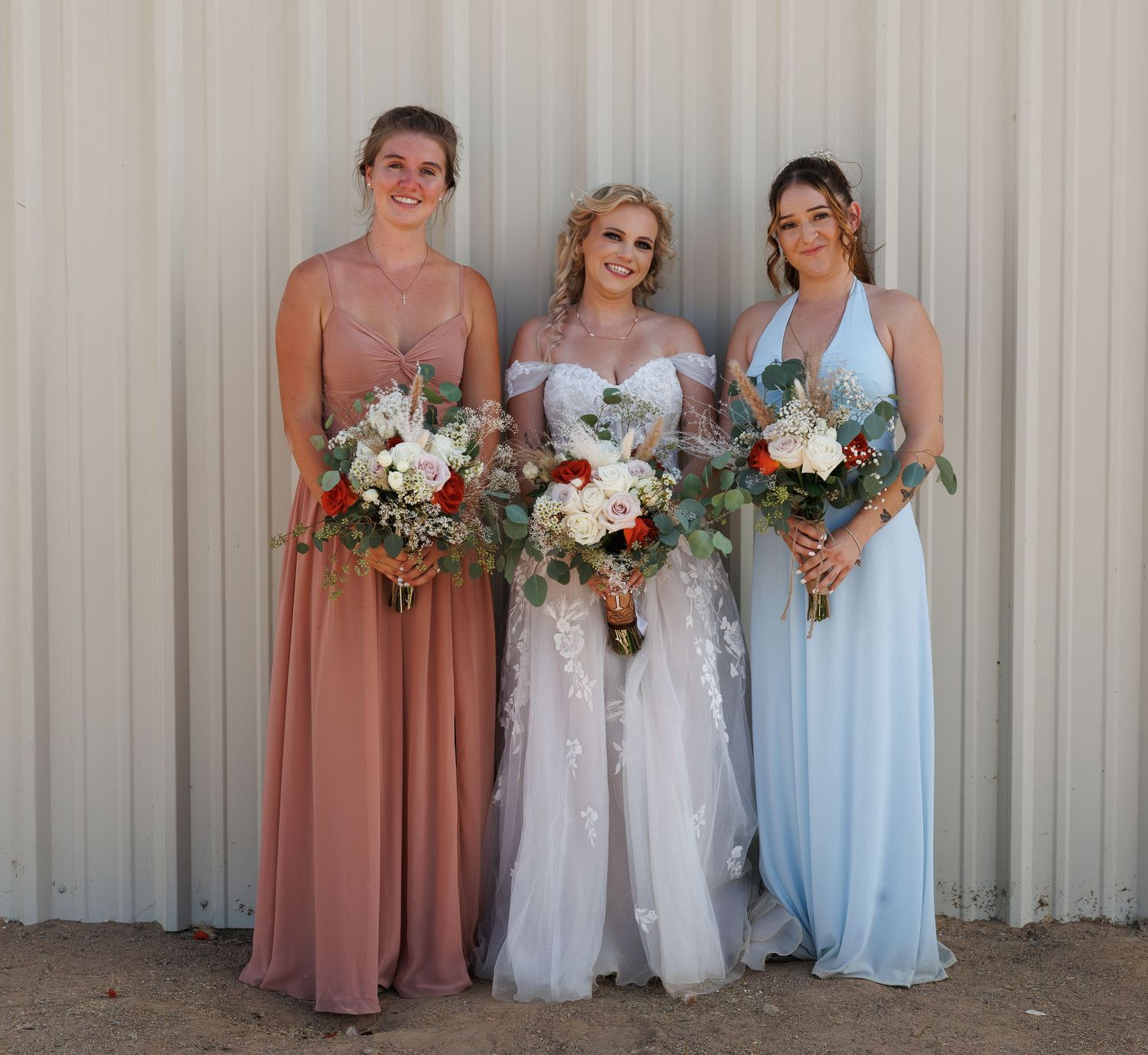 Bride in white lace gown poses with two bridesmaids holding floral bouquets outdoors.