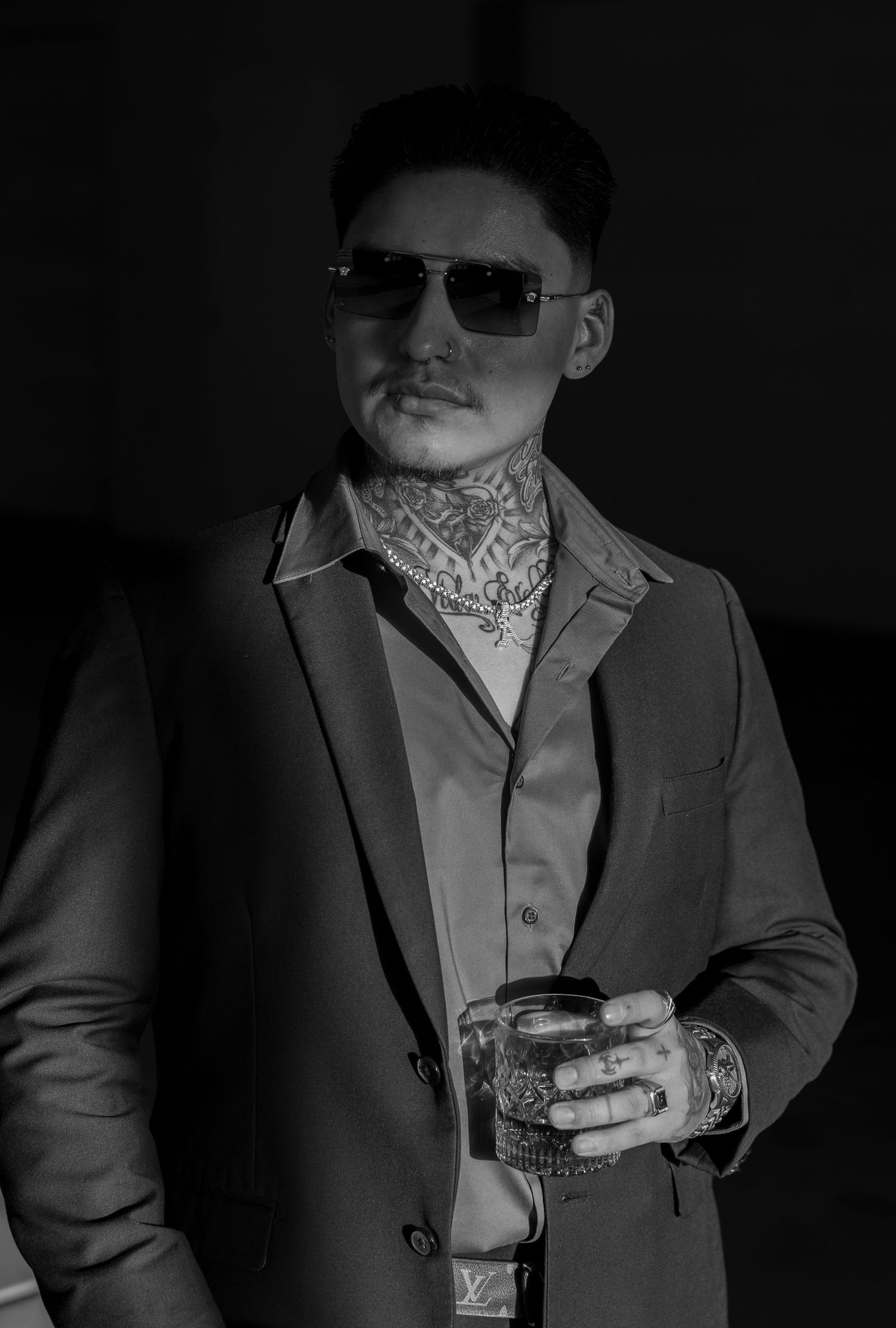 Black and white portrait of tattooed man in suit wearing sunglasses holding a drink with Louis Vuitton bracelet.