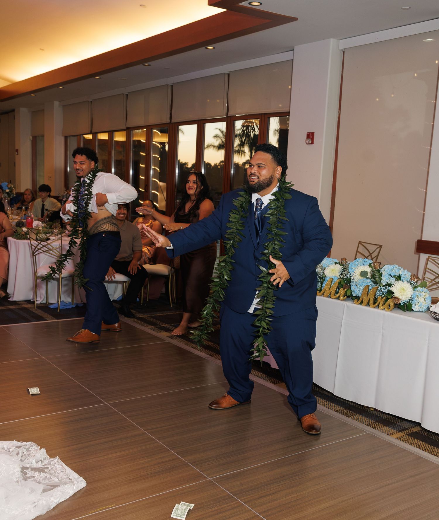 Groom in navy suit and green lei celebrates with guests on dance floor at elegant wedding reception.