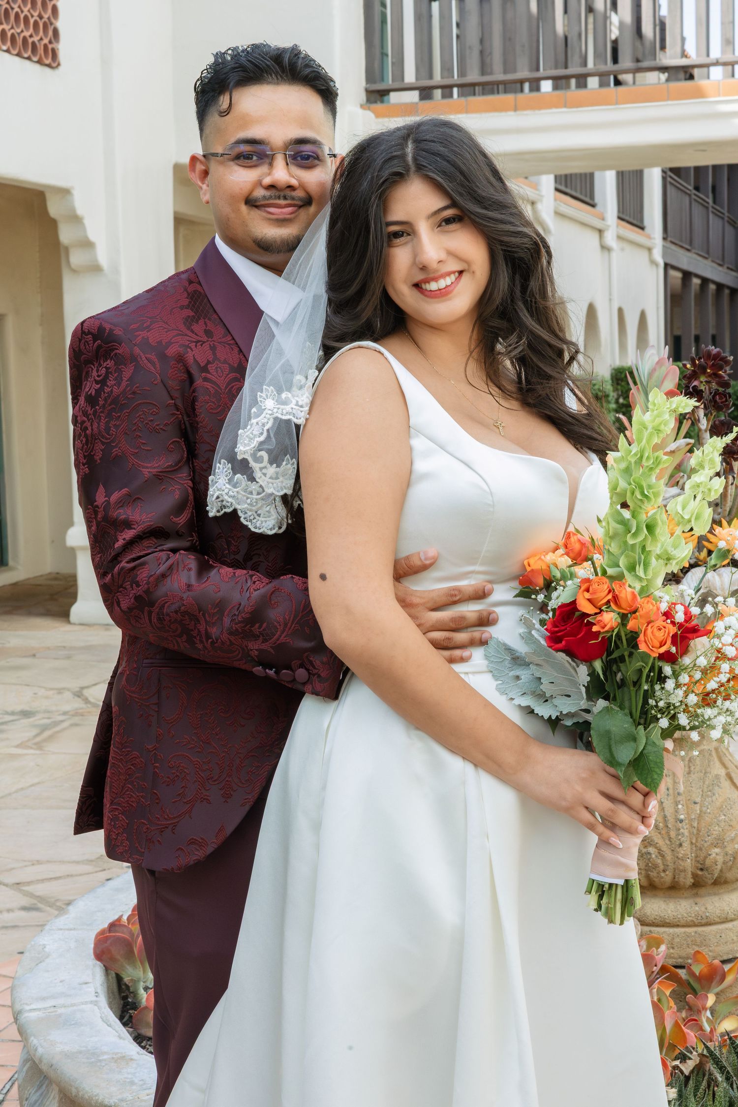 Smiling bride in white dress holding orange bouquet poses with groom in burgundy floral suit outdoors.