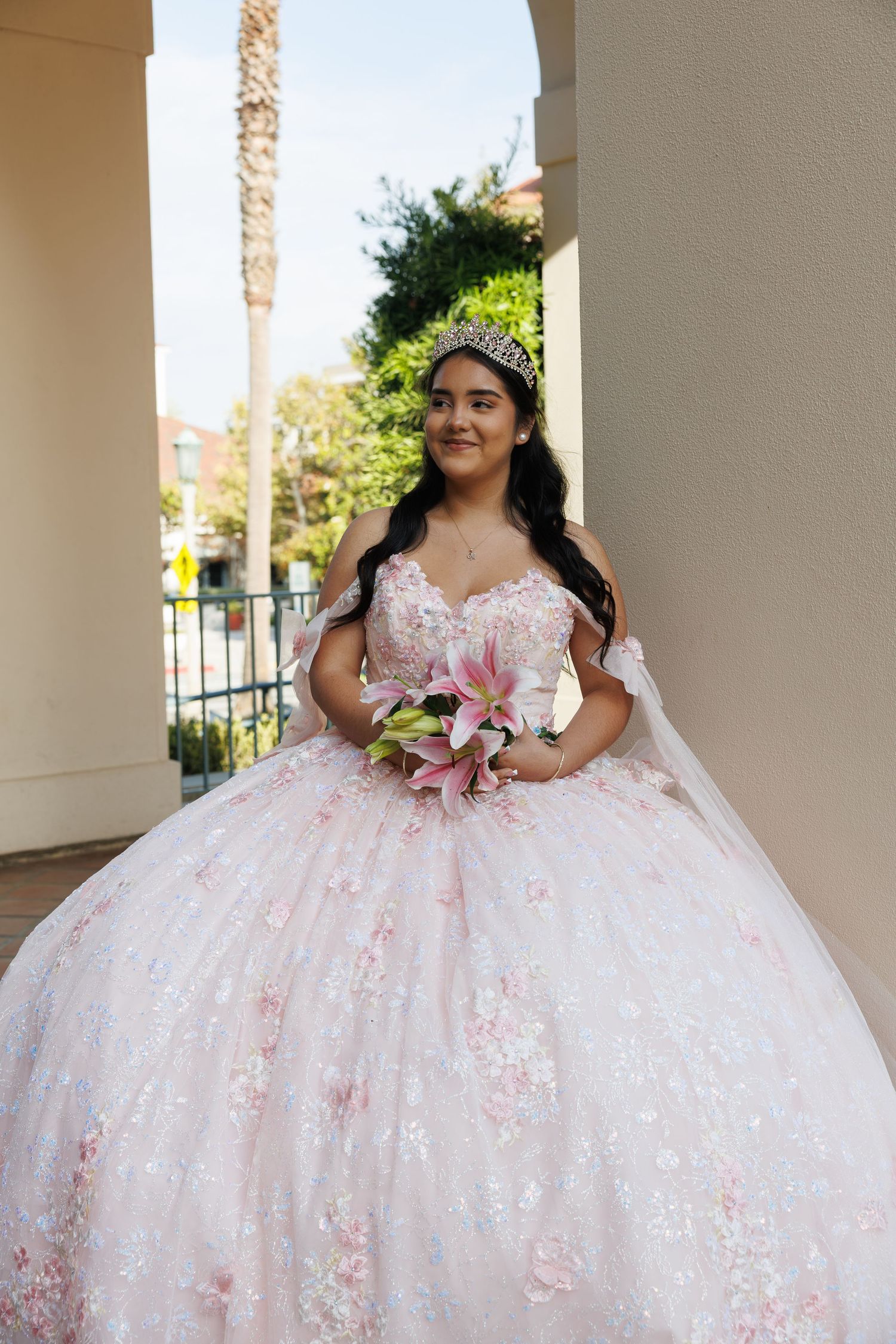 Young woman in pink ball gown and tiara holding floral bouquet during quinceañera celebration outdoors.