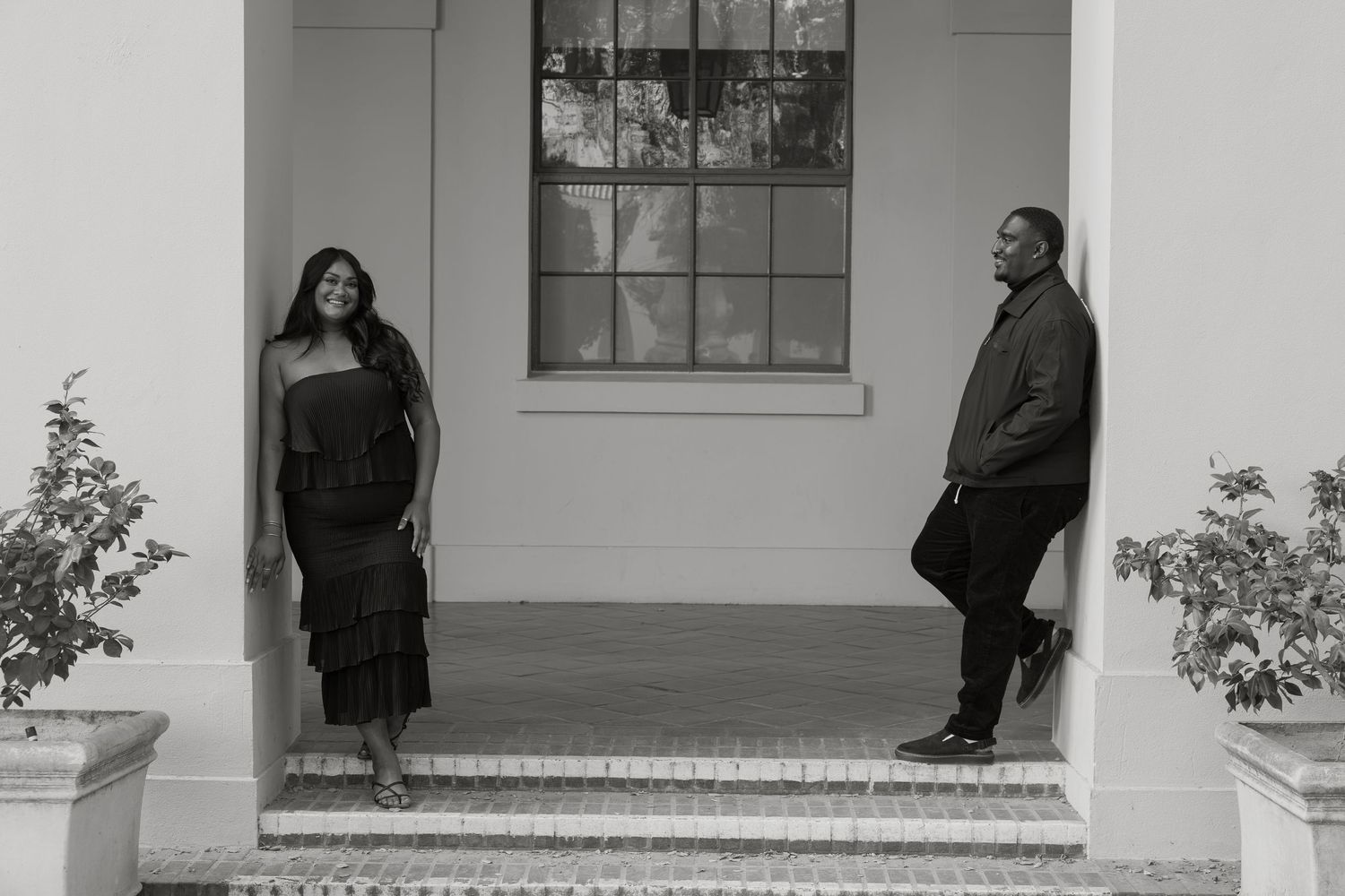 Black and white photo of a couple posing on elegant building steps, woman in ruffled dress, man in suit jacket.