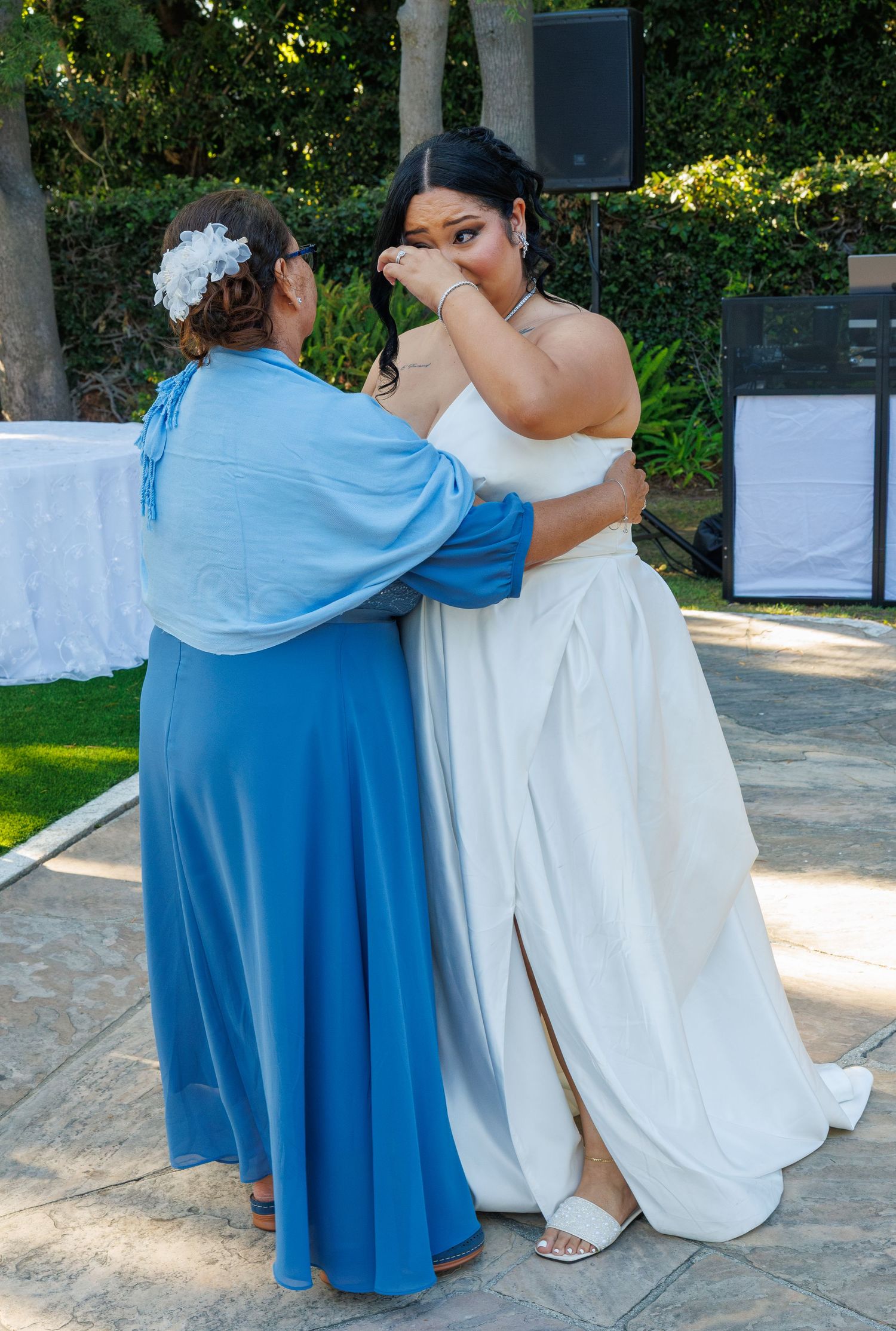 Bride in white gown wipes tears while embracing woman in blue dress at wedding.