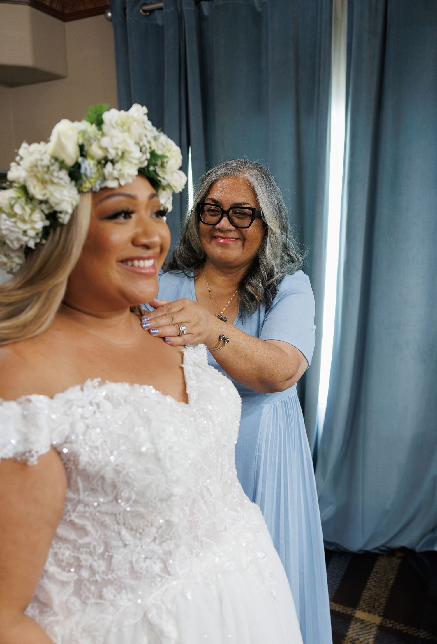 A bride in a white off-shoulder gown and floral crown smiles as a woman in blue adjusts her necklace.