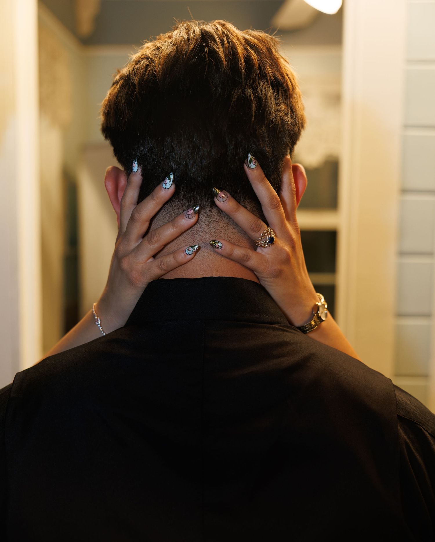 Person seen from behind with hands clasped on neck, wearing black shirt and jewelry, short brown hair.