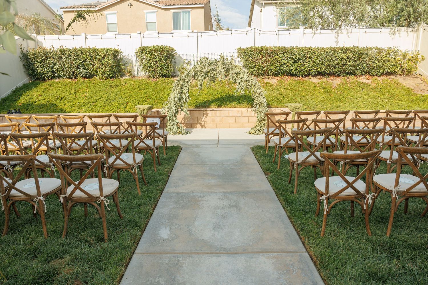 Outdoor wedding ceremony setup with wooden cross-back chairs lining a concrete aisle, green lawn, and floral arch backdrop.