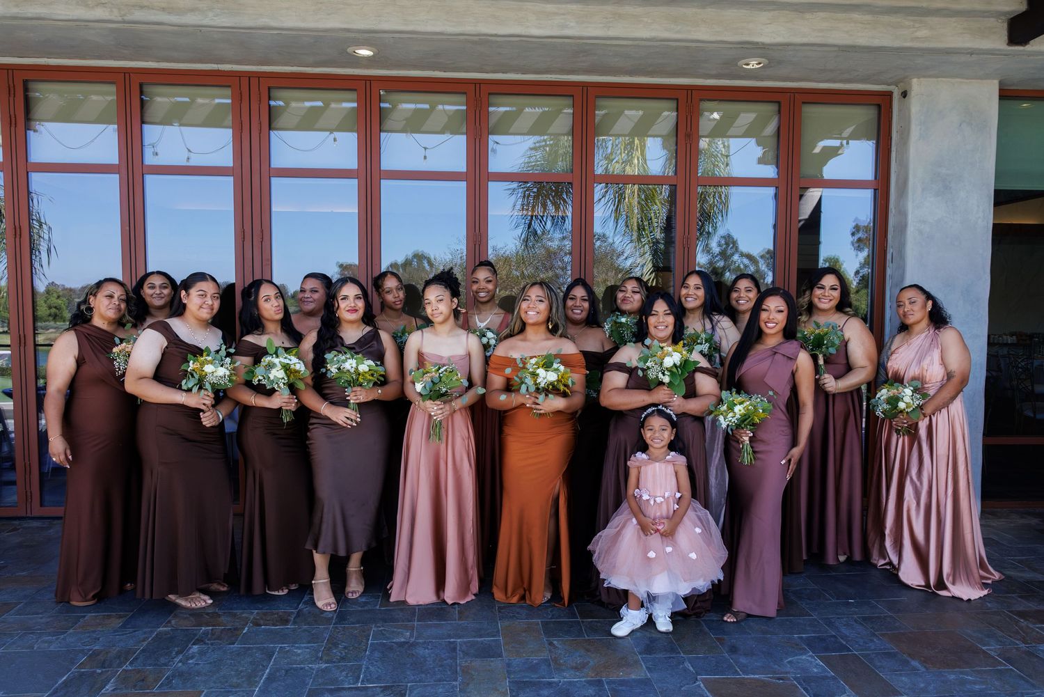 Large bridal party in mismatched brown and mauve gowns posing with bouquets outside elegant venue.