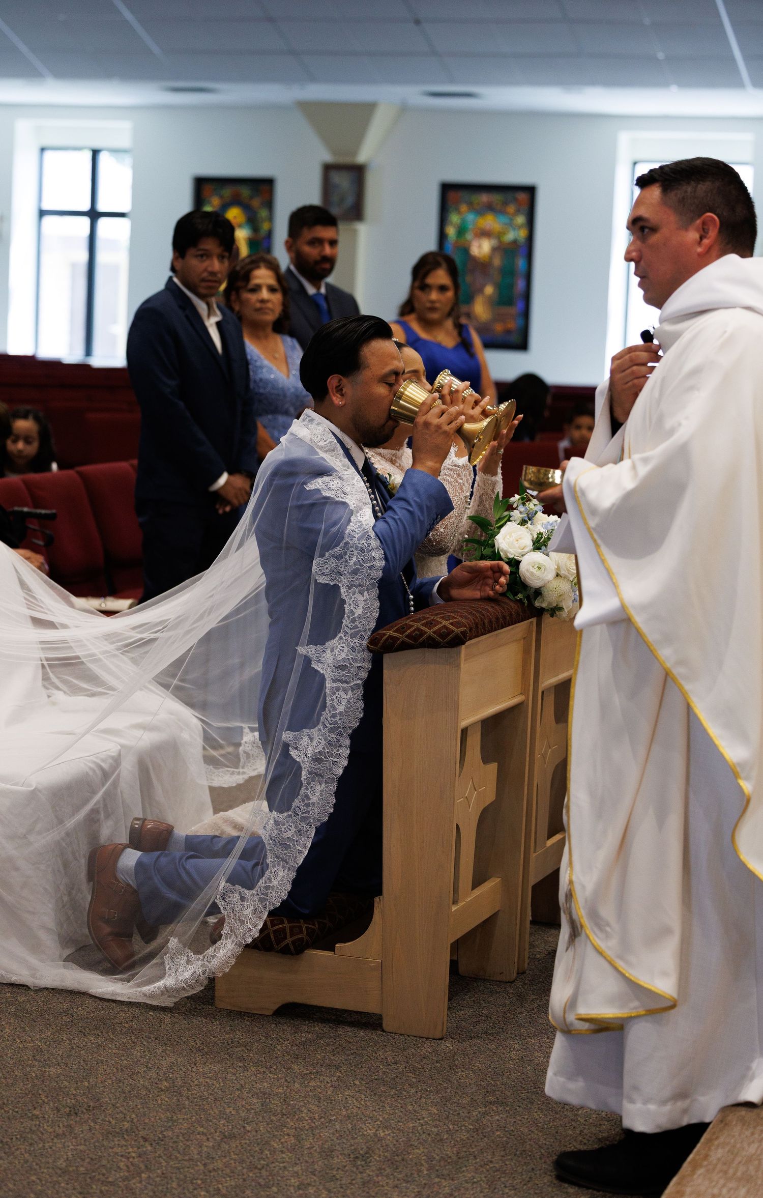 Bride and groom kneel at a church altar during a Catholic wedding Mass as a priest in white vestments offers communion.