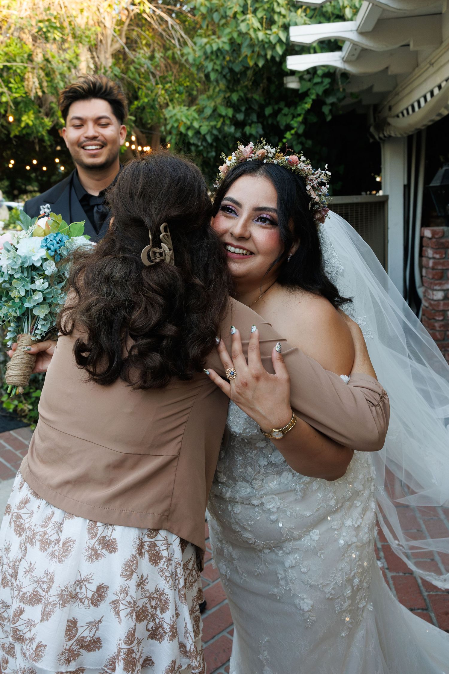 Smiling bride in lace gown and floral crown receives warm hug from wedding guest.