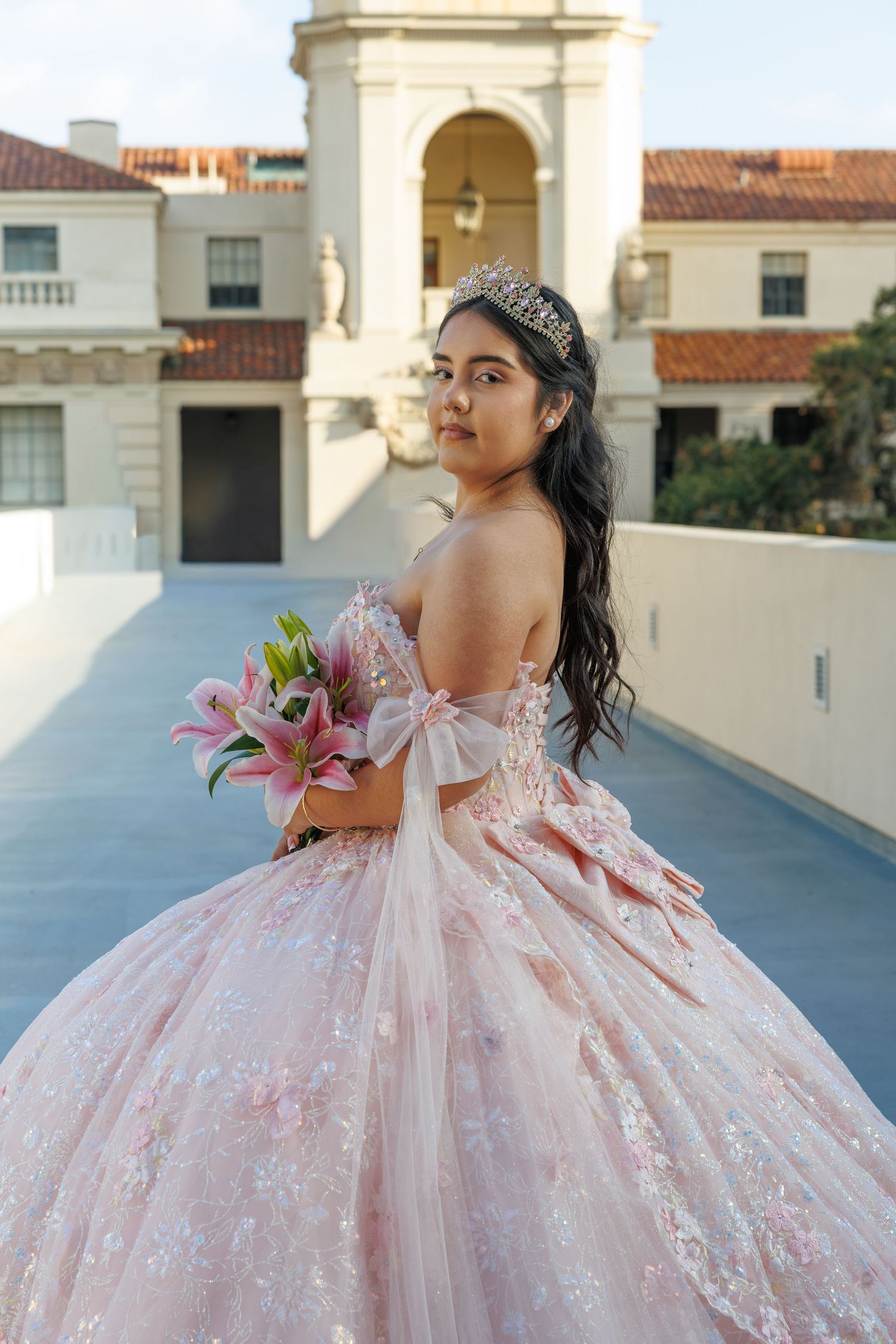 Quinceañera girl in pink strapless ball gown holds pink lilies bouquet outside elegant mansion.