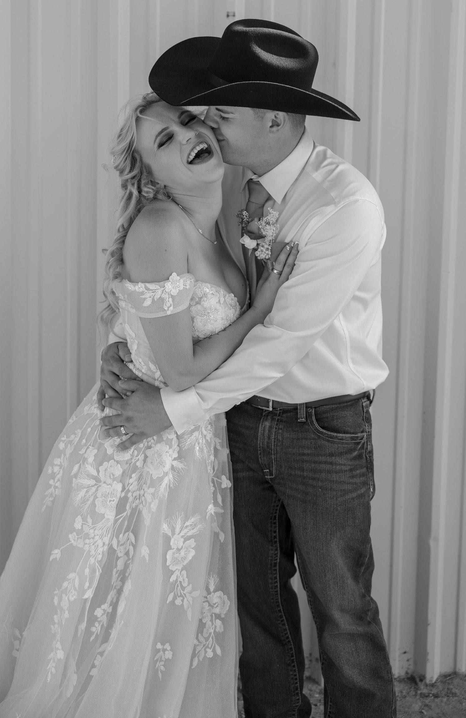 Black and white photo of groom in cowboy hat kissing laughing bride in lace wedding gown.